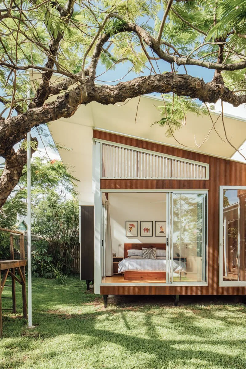 A contemporary elevated house with a sloped roof and wooden facade, featuring an open bedroom through large glass doors, set amidst a sunny, lush garden and framed by a large tree.