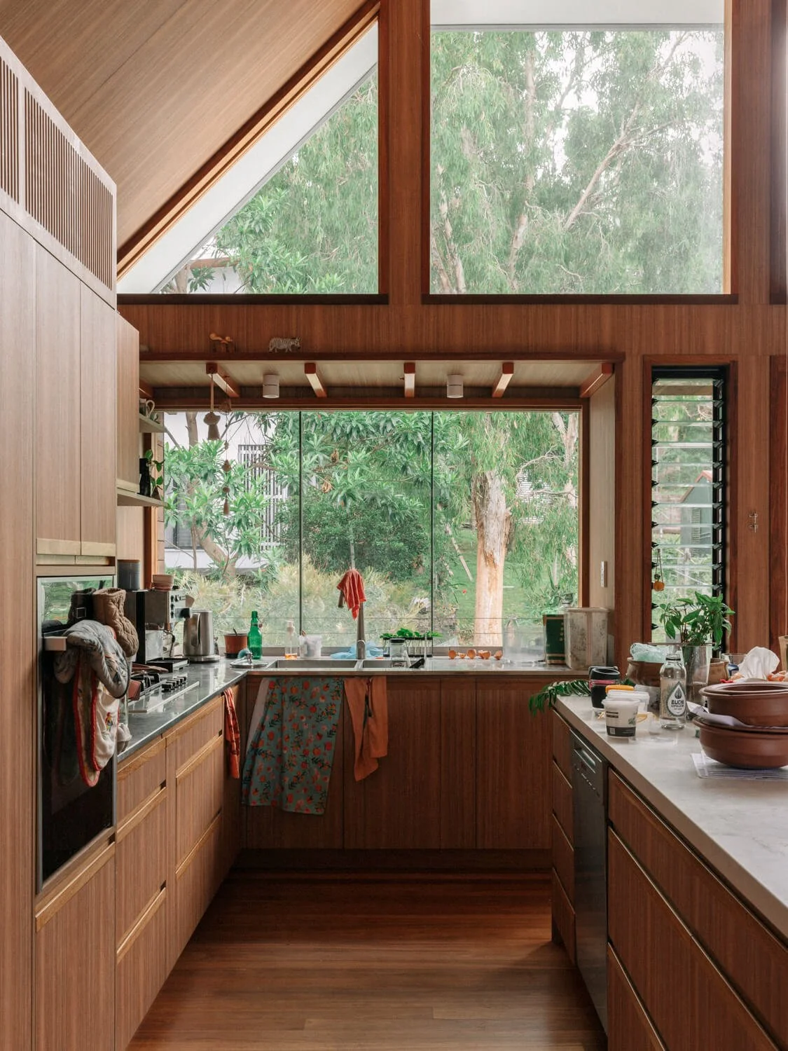 A bright, spacious kitchen features warm wooden cabinetry, an A-frame ceiling, and large windows that look out onto a vibrant green tree-filled landscape.