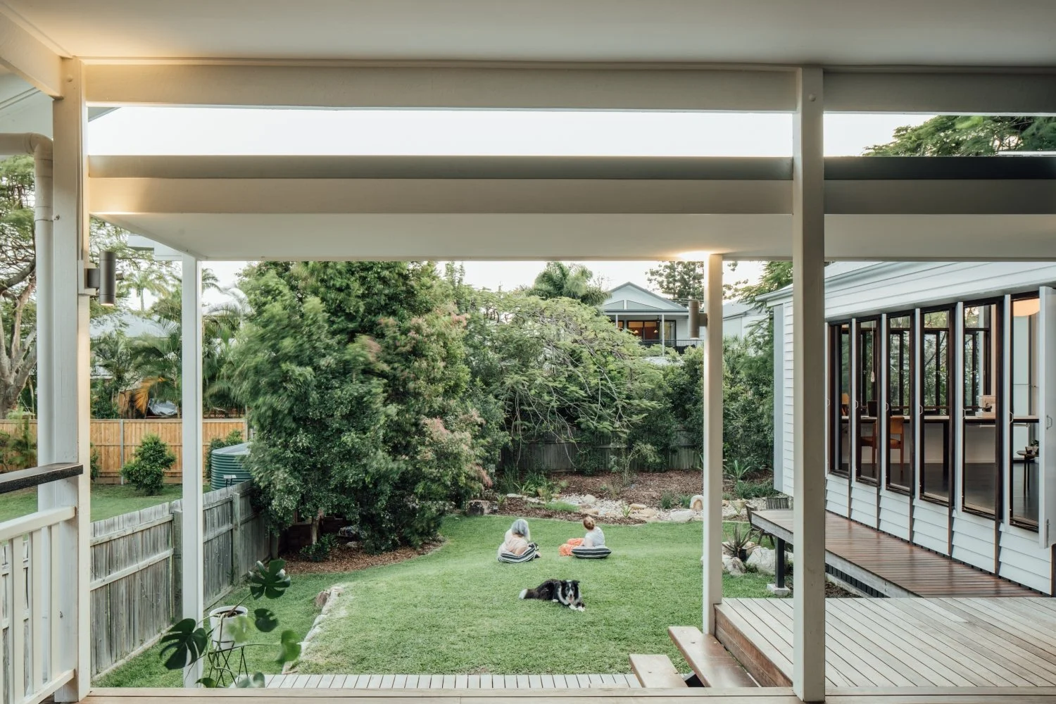 From a modern covered deck, two children and a dog relax on a verdant backyard lawn, bordered by trees and a house with wide open glass doors.