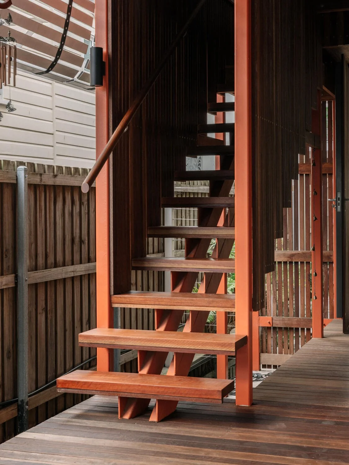 A modern outdoor staircase with open wooden treads and reddish-orange supports rises from a dark wooden deck, flanked by vertical wooden screens and a white-sided building in the background.