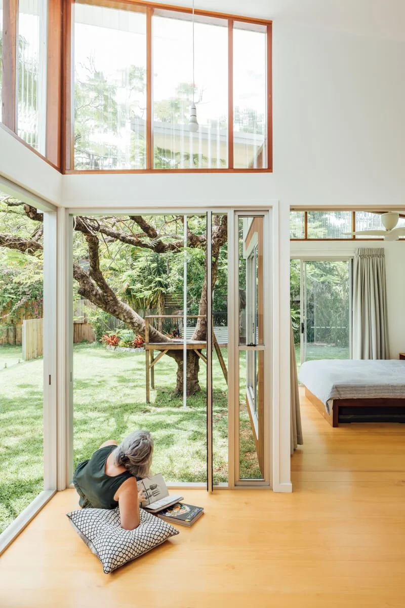A person reads a book on a cushion by expansive windows, connecting a bright modern room to a lush backyard with a treehouse and an adjacent bedroom.