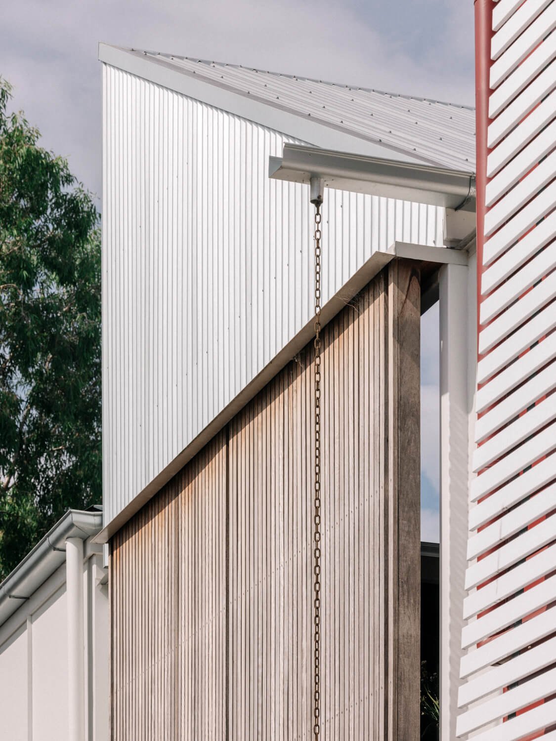 Modern building exterior combining corrugated metal, weathered vertical wood siding with a rain chain, and a white slatted screen with a red column, set against a sky and green trees.