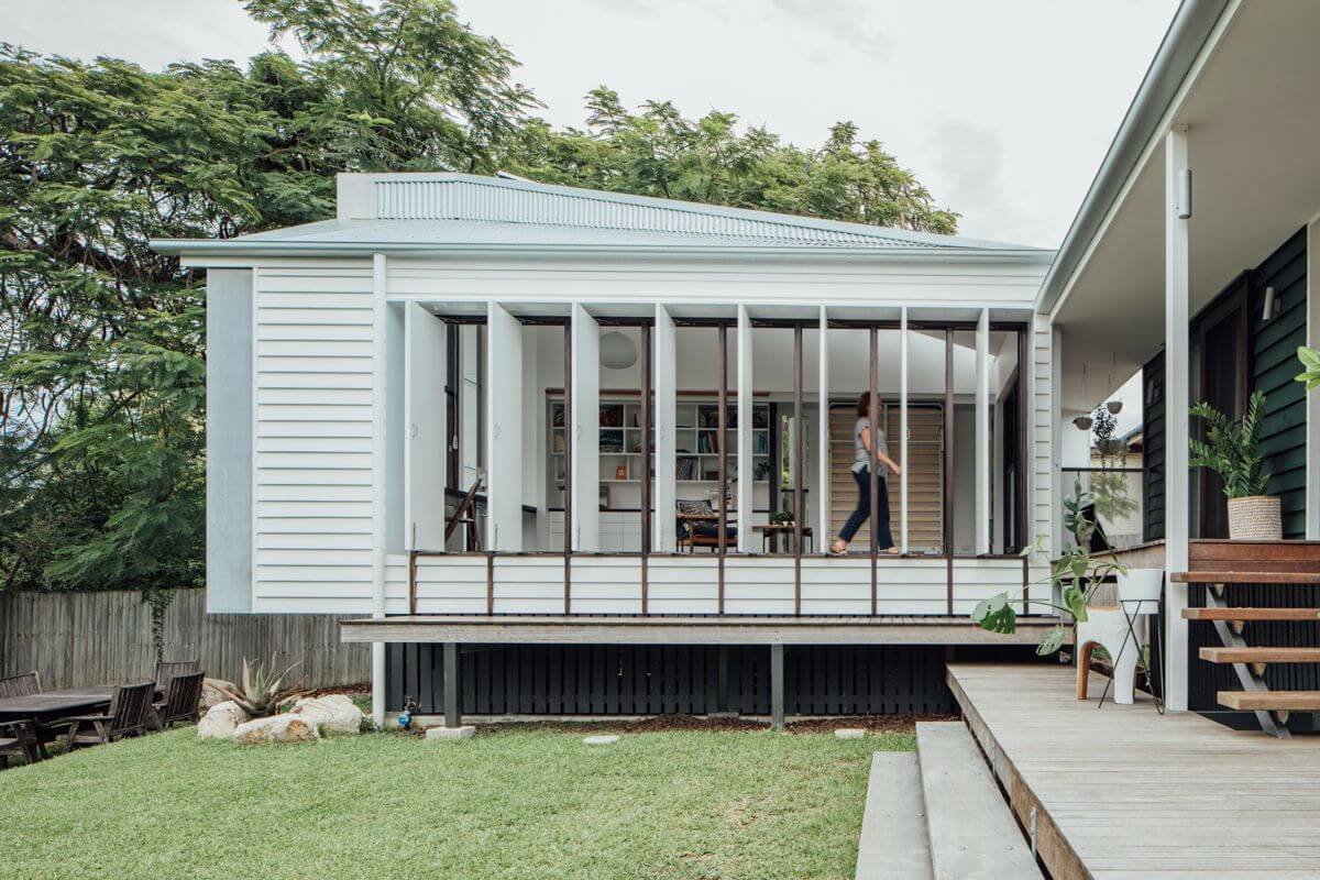 A woman walks through a bright, elevated room with white folding window panels, part of a contemporary house with a connected wooden deck overlooking a green yard and tall trees.