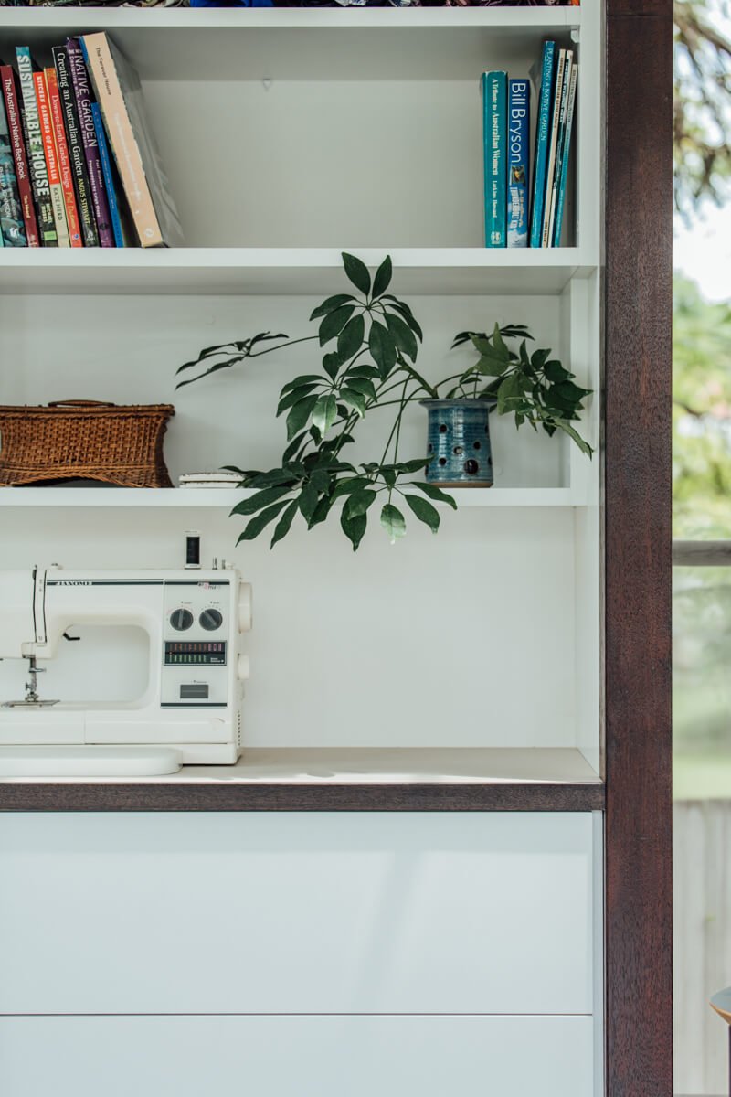 A modern white shelving unit with dark wood trim holds a white sewing machine on its counter, with books, a potted plant, and a woven basket on shelves above, beside a window revealing outdoor greenery.