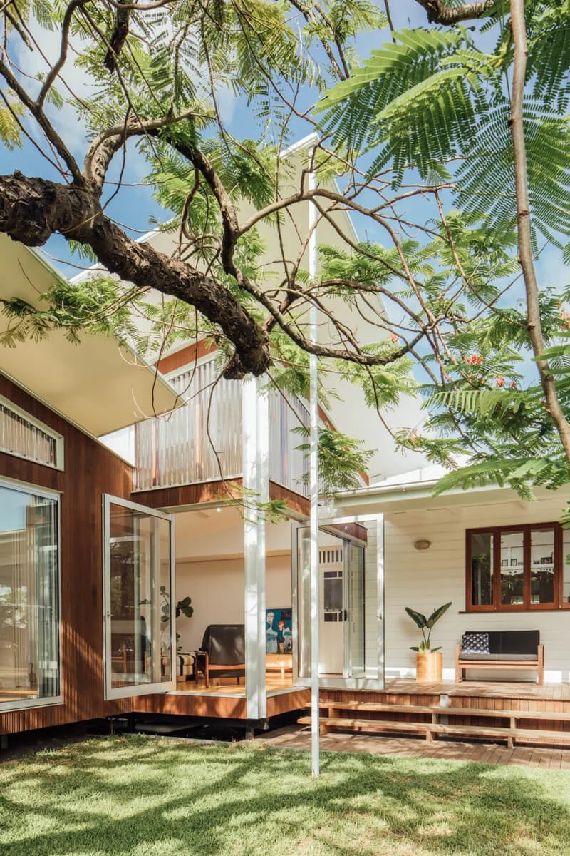 A contemporary home with warm wood and white paneling features open glass doors leading to a wooden deck and grassy lawn, beneath the canopy of a large, leafy tree.