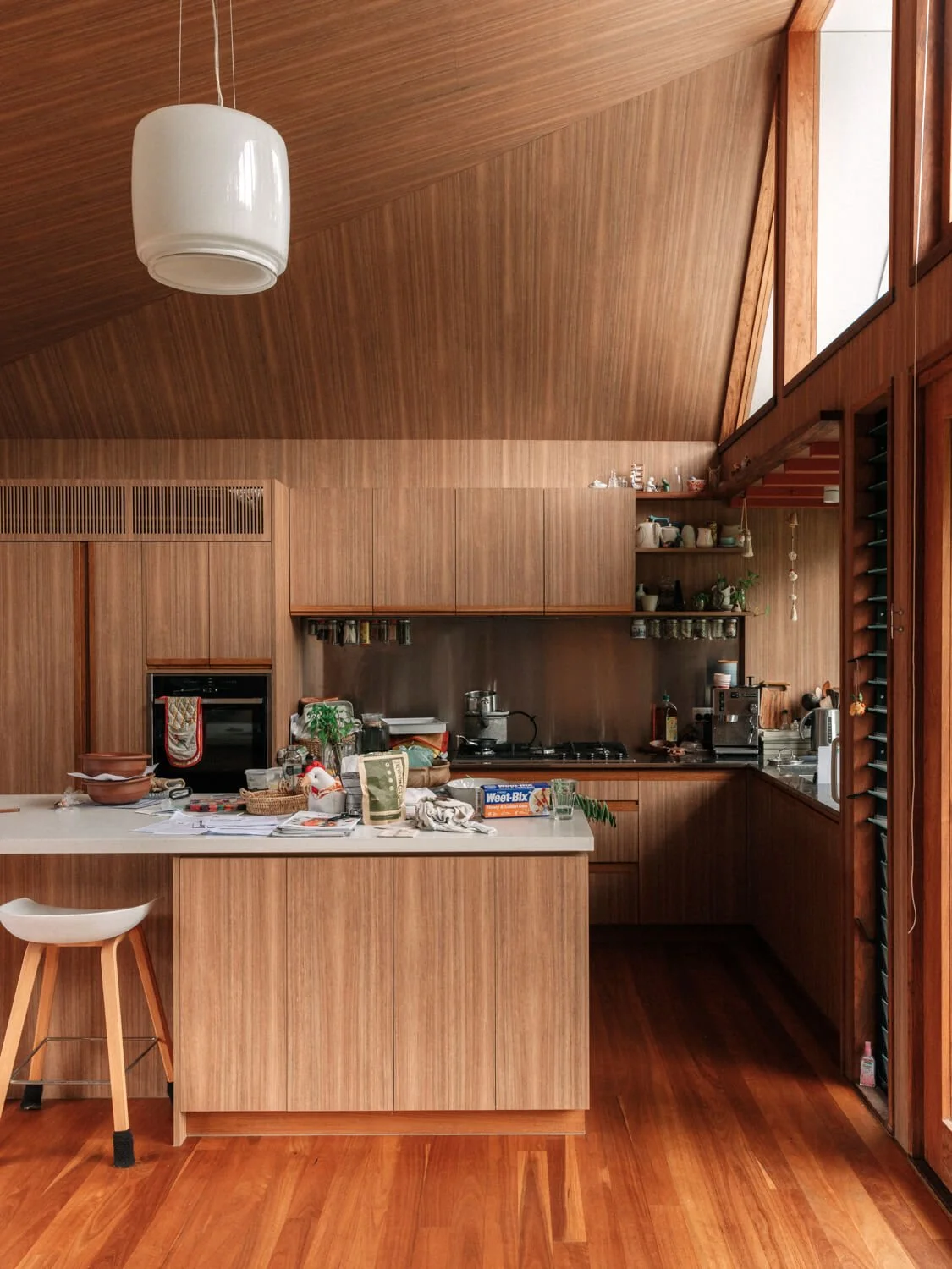 A warm, modern kitchen features extensive wood paneling across its floor, cabinets, and dramatic vaulted ceiling, with an island counter holding everyday items and bright natural light.