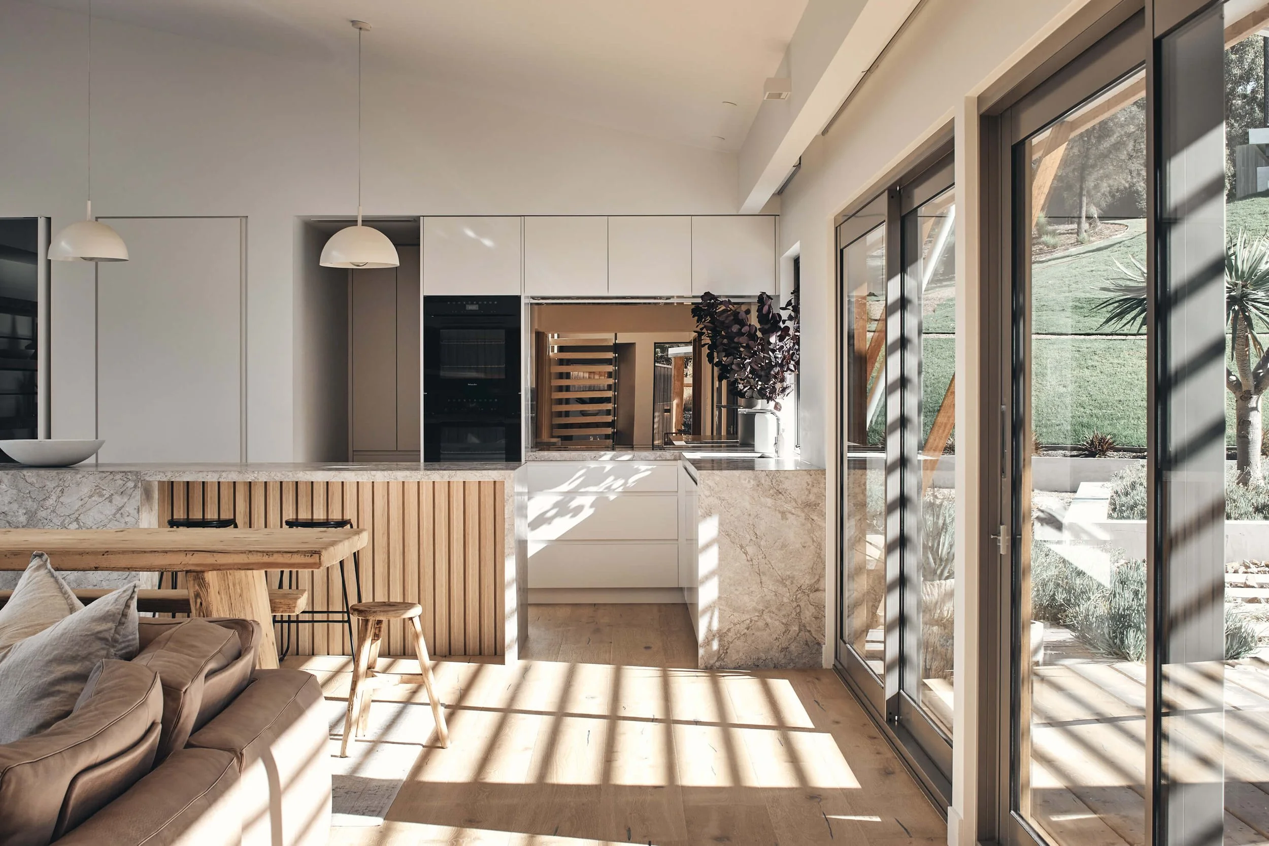 A sunlit, modern open-plan kitchen and living area features a wooden dining table, a marble-topped island, and large glass doors opening to a lush green outdoor space.
