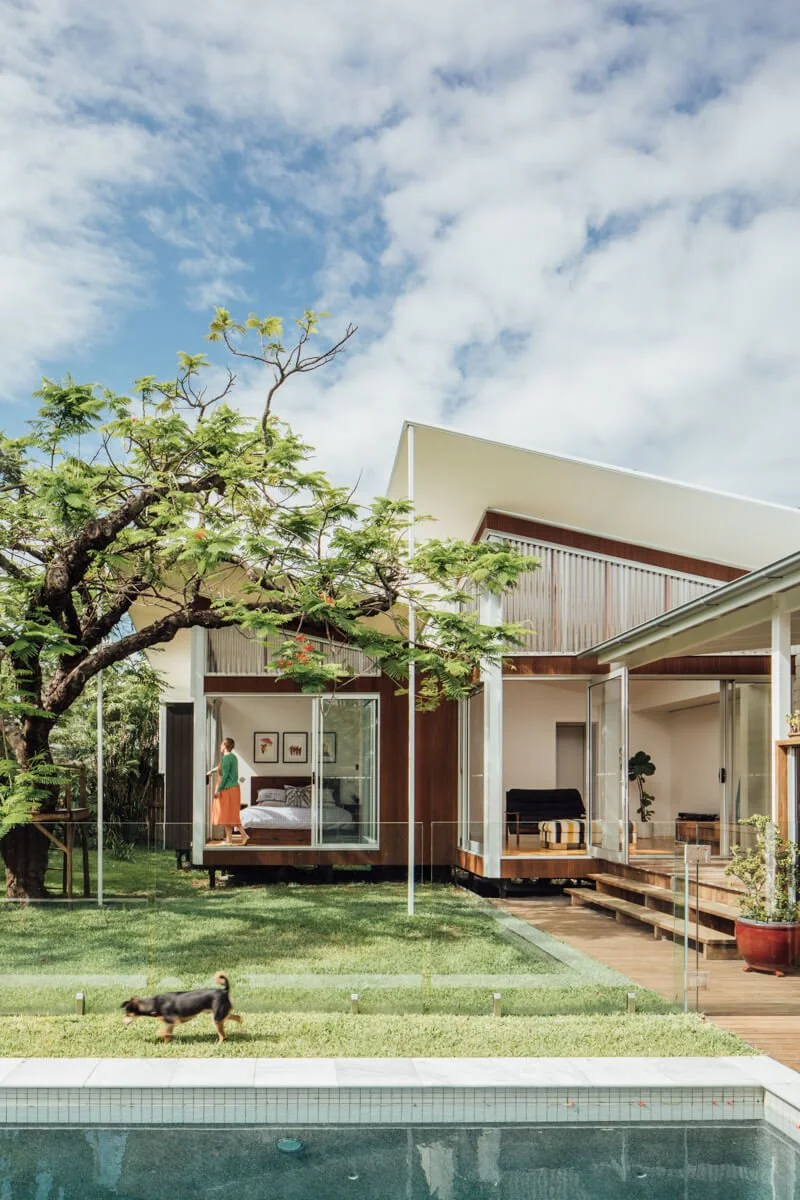 A modern house with large glass windows and an angled white roof overlooks a green lawn where a dog runs towards a swimming pool, while a woman looks out from a bedroom and a large tree frames the left side under a bright, cloudy sky.