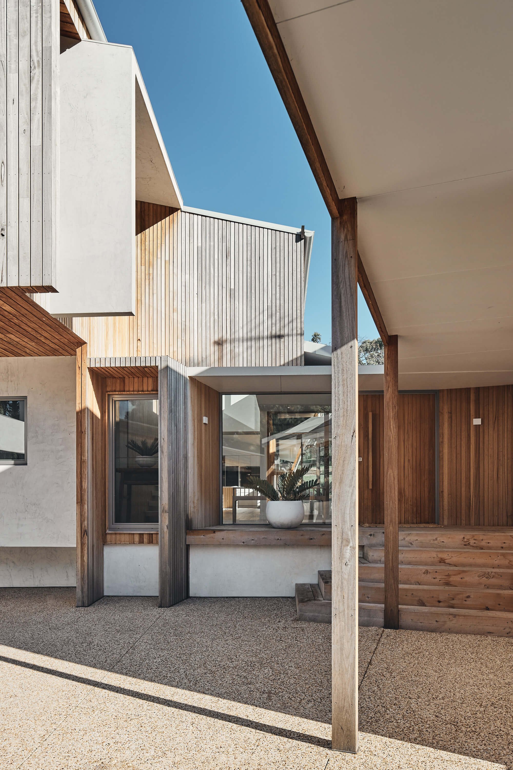 A contemporary house exterior showcases a harmonious blend of light wood paneling, crisp white stucco, and large glass windows, all under a bright blue sky with strong sunlight casting geometric shadows on the speckled aggregate ground.