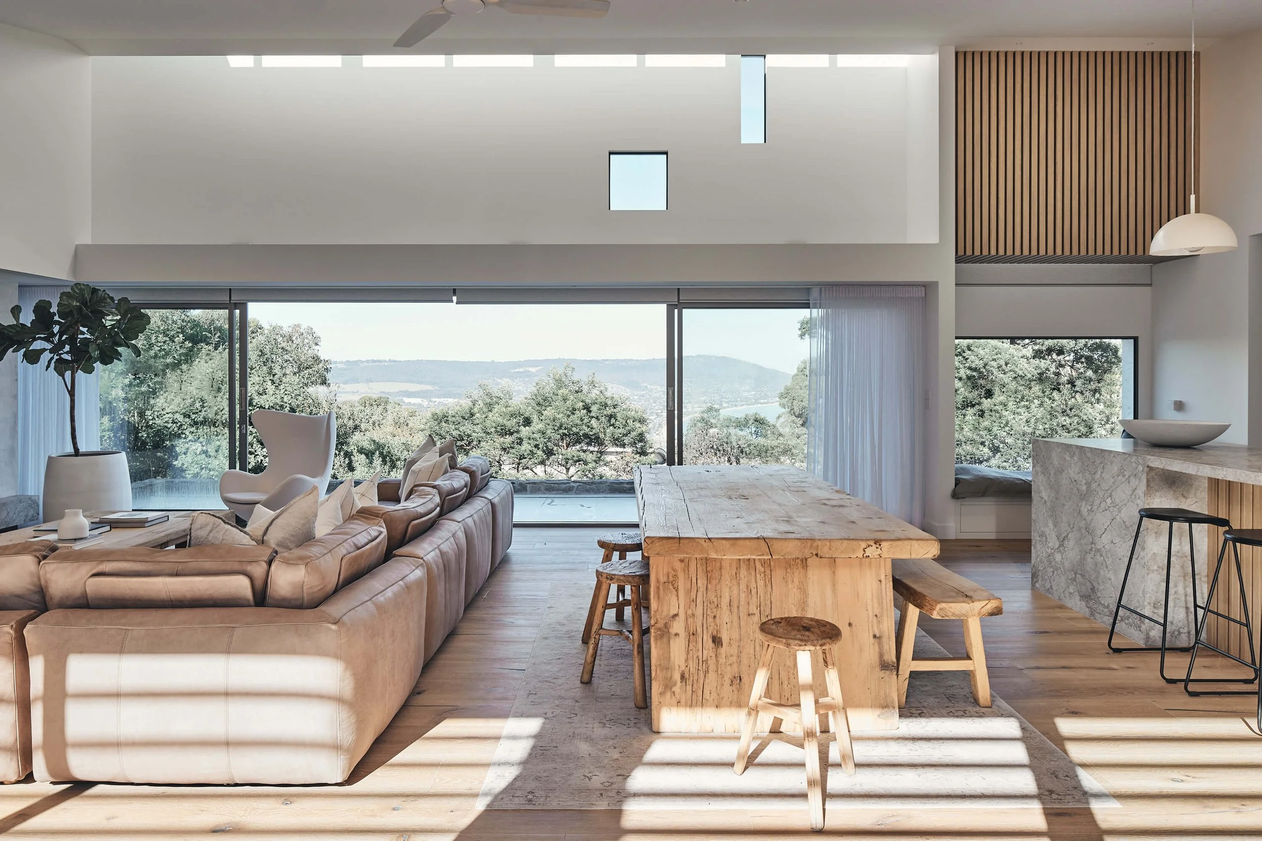 A bright, open-concept living and dining space features a tan leather sectional sofa, a rustic wooden table, and a marble kitchen island, all framed by expansive windows offering a panoramic view of a lush, hilly landscape.