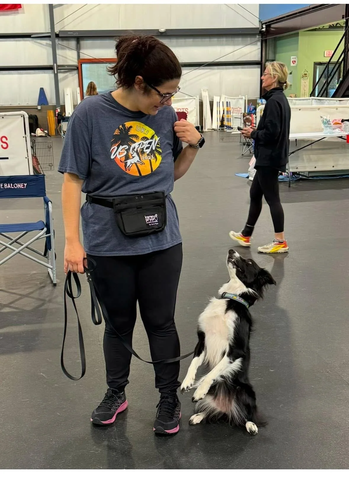 A woman in athletic clothing and glasses playing with a black and white border collie puppy indoors at a dog training or event facility. The puppy is sitting on its hind legs and looking up at her.