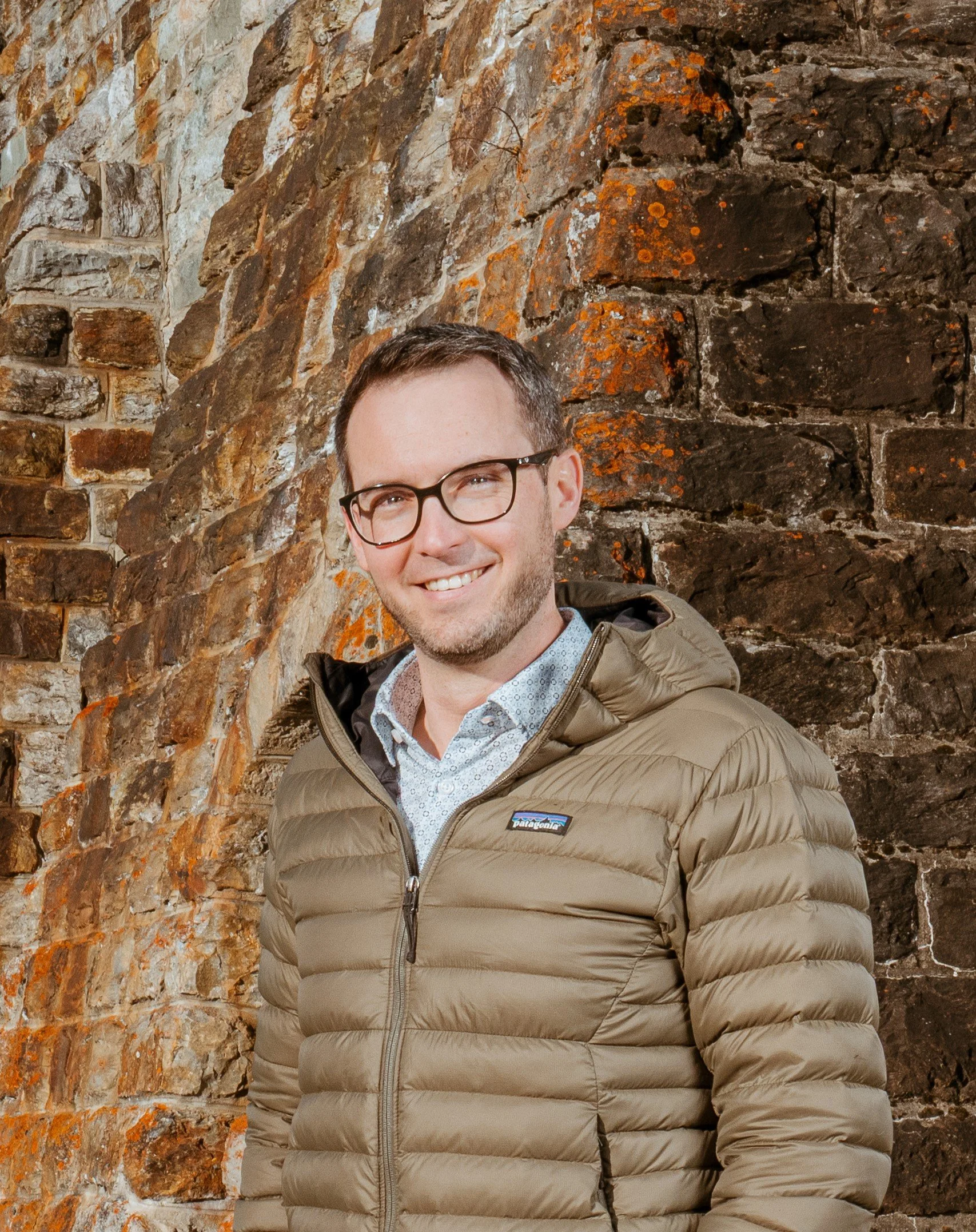 Ein lachender Mann mit Brille trägt eine beige Daunenjacke und steht vor einer alten rostigen Backsteinmauer.