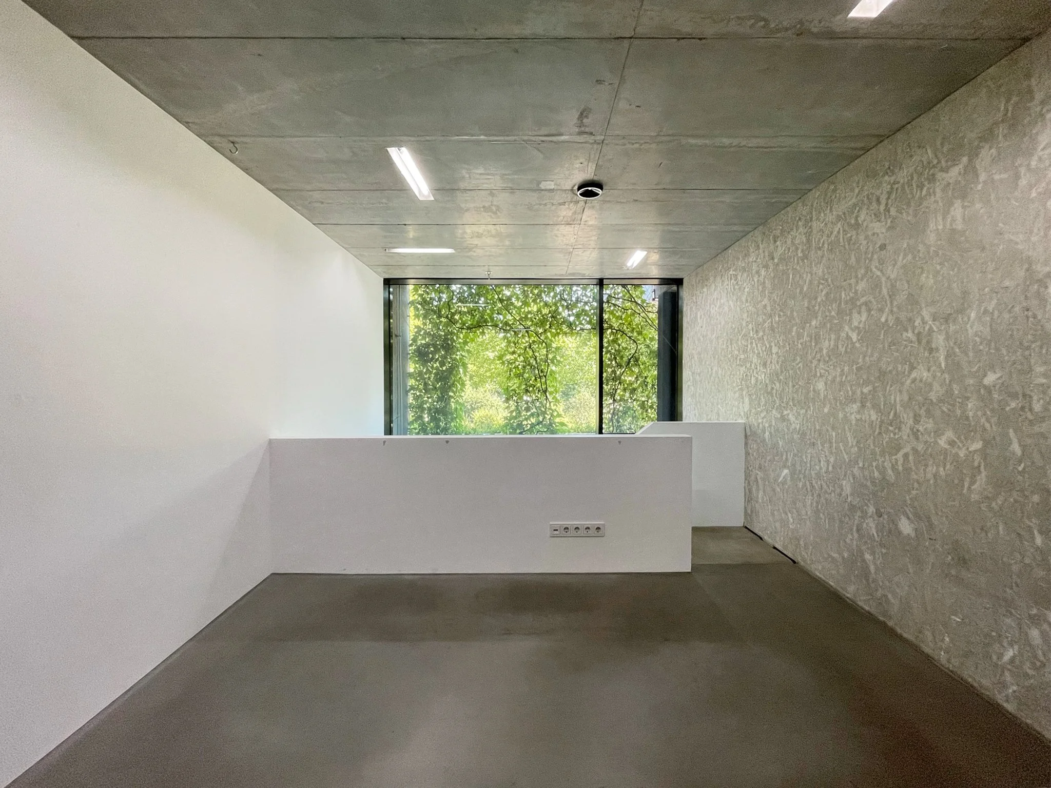 Ein moderner leerer Raum mit großen Fenstern, weißen und grauen Wänden sowie verputzten Böden, vermutlich in einem Bürogebäude oder einer Galerie.