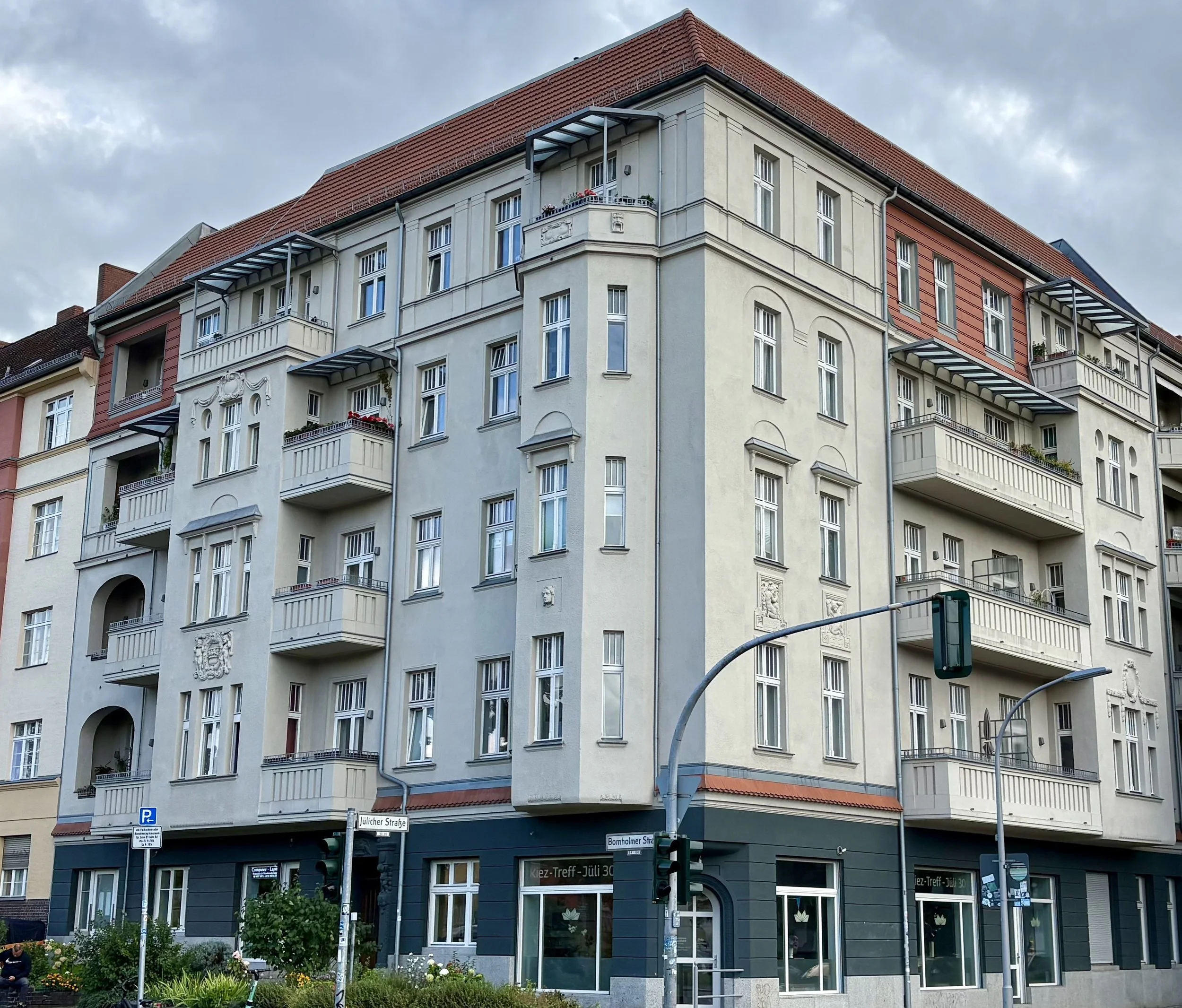 Mehrstöckiges Wohngebäude mit Balkonfenstern, Balkonen und einer roten Dachgaube, an einer Straßenecke in einer Stadt, mit Straßenschildern und Straßenlaternen vorn.