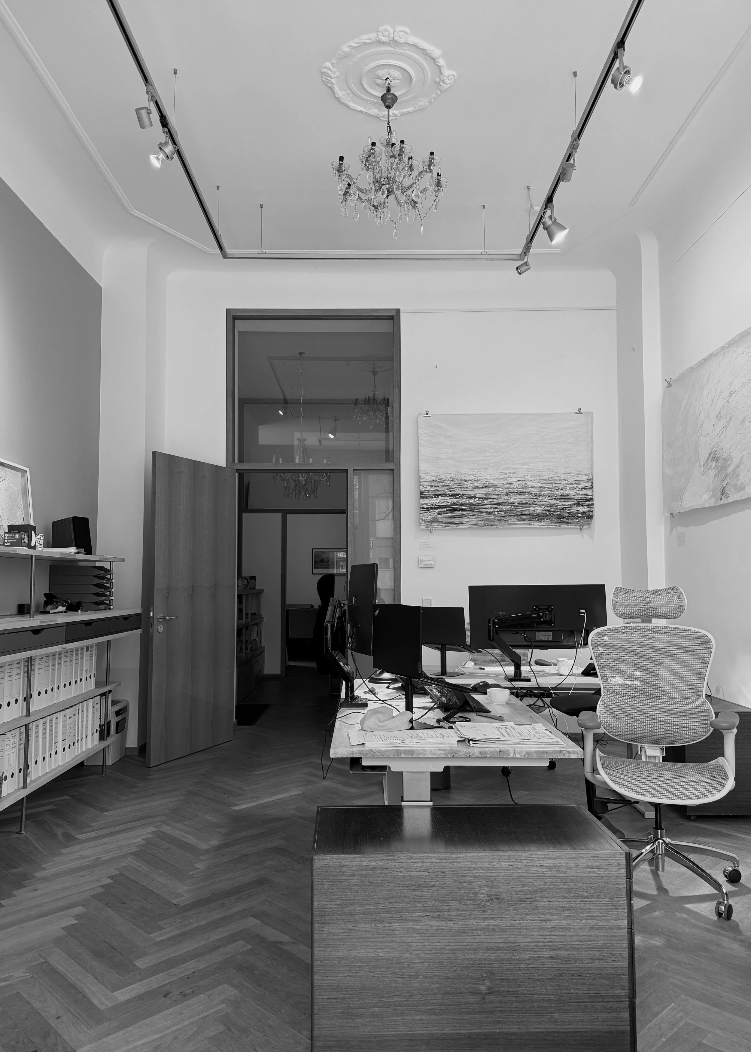 Ein modernes Büro mit mehreren Computerbildschirmen, einem weißen Bürodrehstuhl und einem großen Tisch mit Unterlagen. Im Hintergrund hängen zwei Gemälde an der Wand, der Raum hat eine hohe Decke mit Kronleuchter und Deckenleuchten.