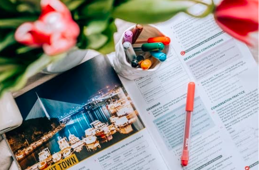 Une table avec un magazine ouvert, des crayons de différentes couleurs dans un bol, un stylo à encre rose, et un bouquet de fleurs roses et vertes.