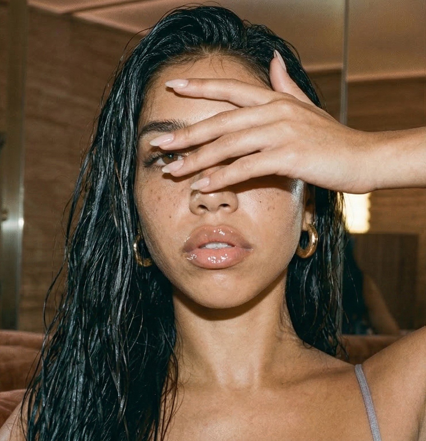 Close-up of a woman with wet dark hair, applying a moisturized, glossy look to her lips, with her left hand covering part of her face, showing gold hoop earrings and a gray spaghetti strap top.