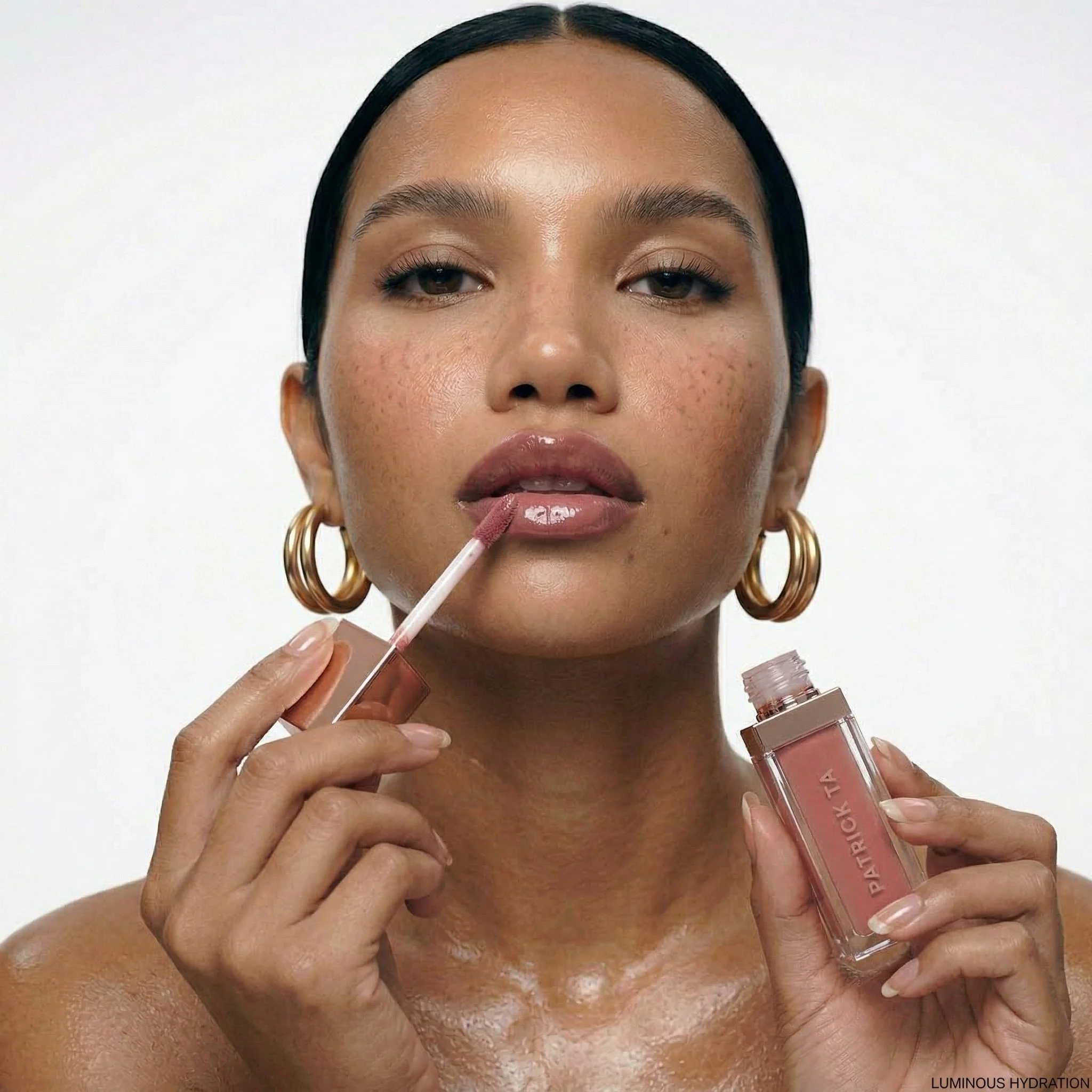 A woman with dark hair and gold hoop earrings applies nude lip gloss from a Patrick Ta lip gloss tube, holding a wand near her lips.
