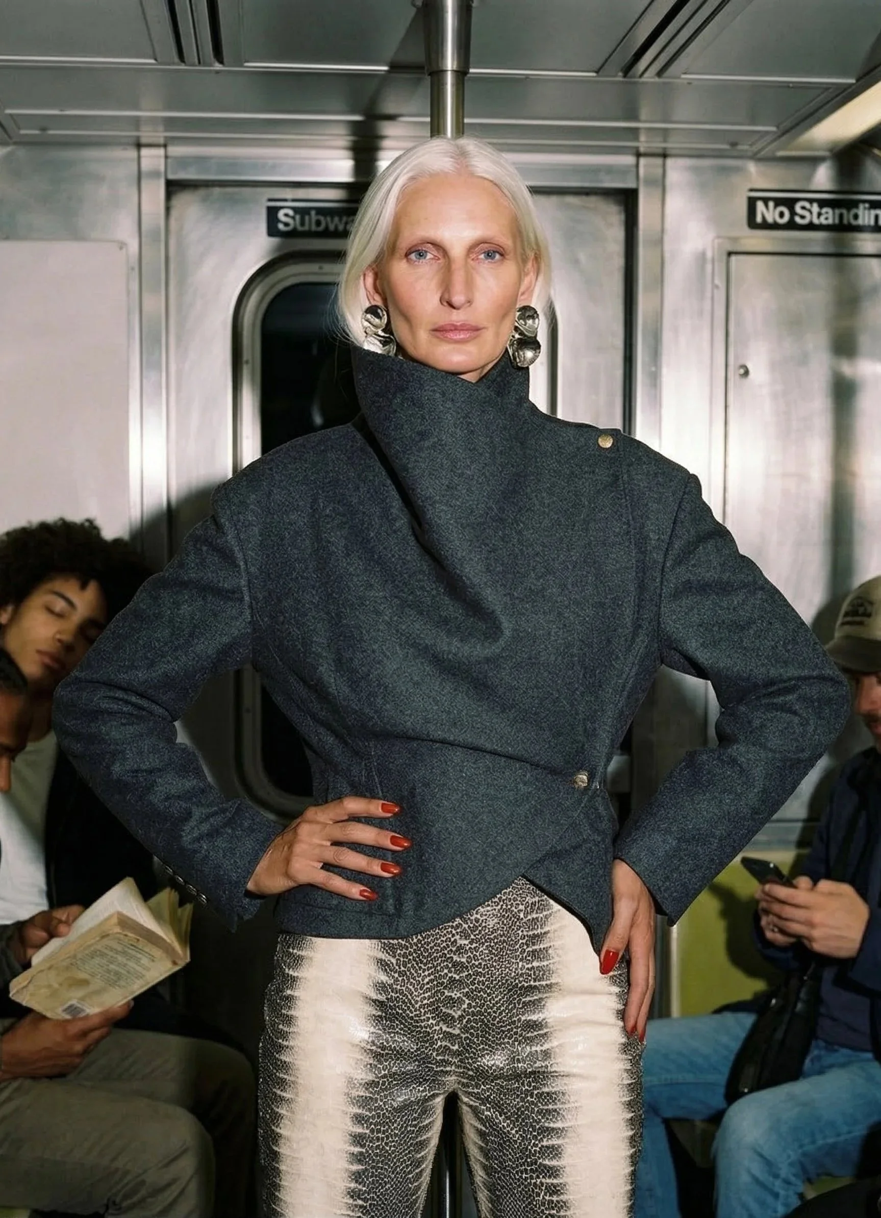 A fashionable woman with platinum blonde hair standing in a subway car, wearing a dark gray high-collared coat and shiny patterned pants, with her hand on her hip and large metallic earrings.