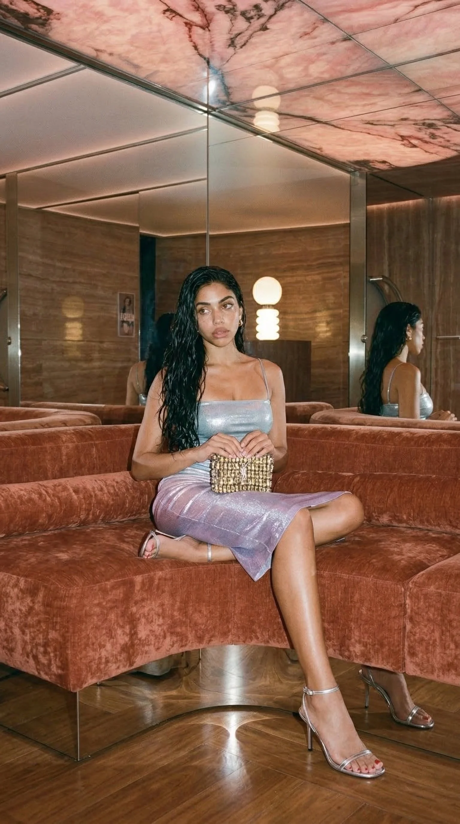 A woman with long black wet hair sitting on a velvet orange sofa in a stylish room with wooden walls and mirrors. She is wearing a shiny silver dress, silver high heels, and holding a small gold purse.