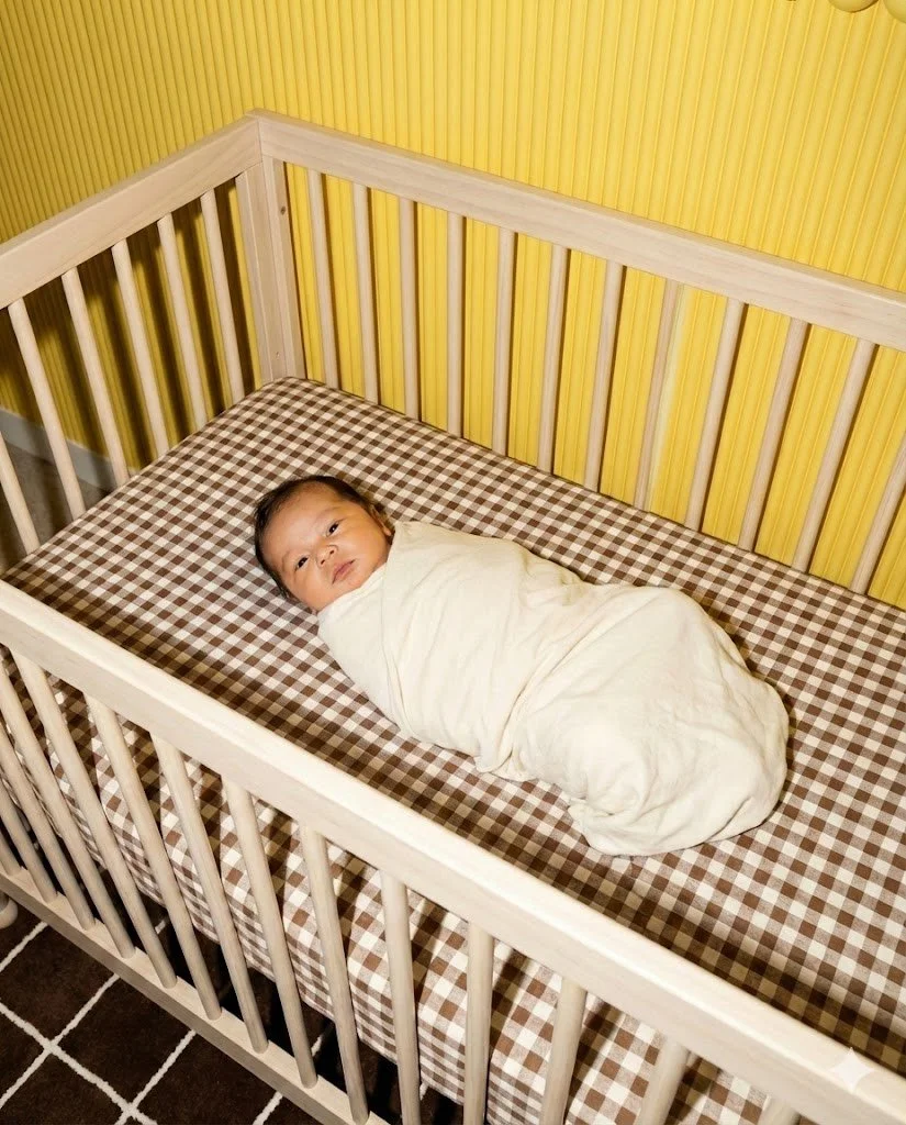 A baby wrapped in a white blanket lying in a crib with a brown and white checkered sheet, in a room with yellow wooden panel walls.
