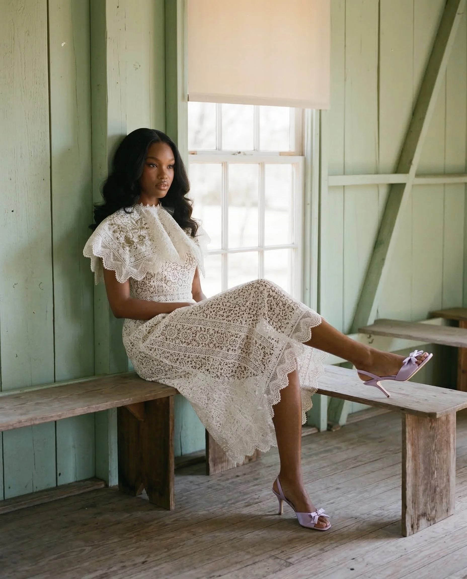 A woman in a white lace dress and purple heels sitting on a wooden bench inside a rustic, green-walled room with large window. Studio Abdul ai campaign image 