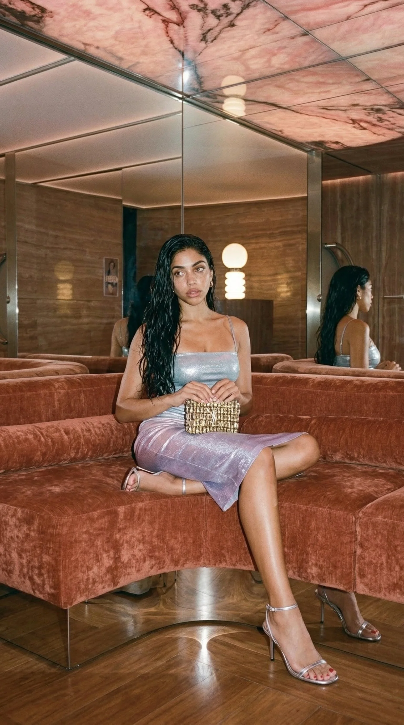 A woman with long dark wavy hair sitting on a rust-colored velvet couch in front of a mirror, holding a gold clutch, dressed in a shimmery silver dress and clear high heels, in a room with wooden walls and modern lighting.