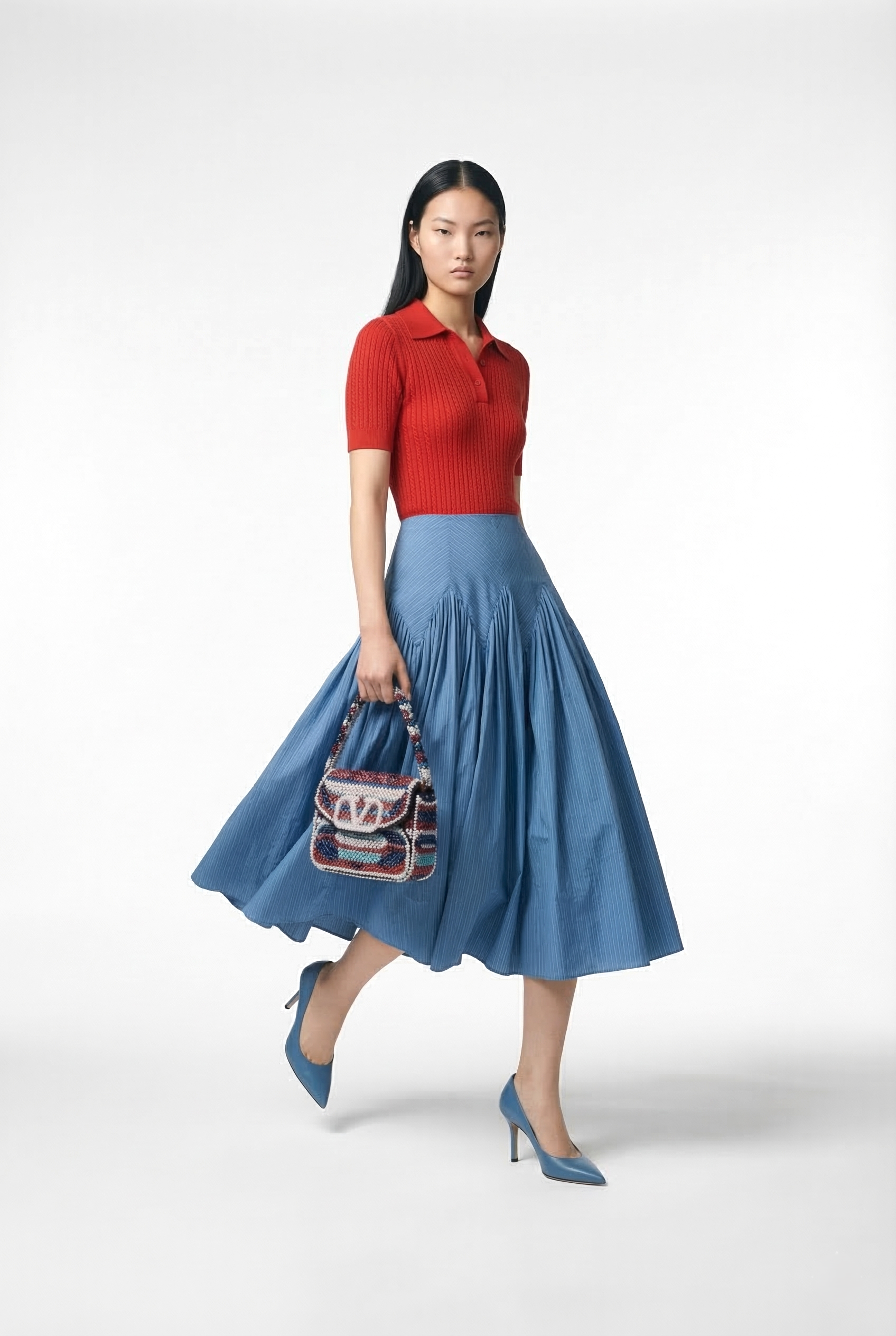 A woman walking wearing a red short-sleeve polo shirt, a blue pleated midi skirt, matching blue high heels, and carrying a colorful woven handbag against a plain white background.