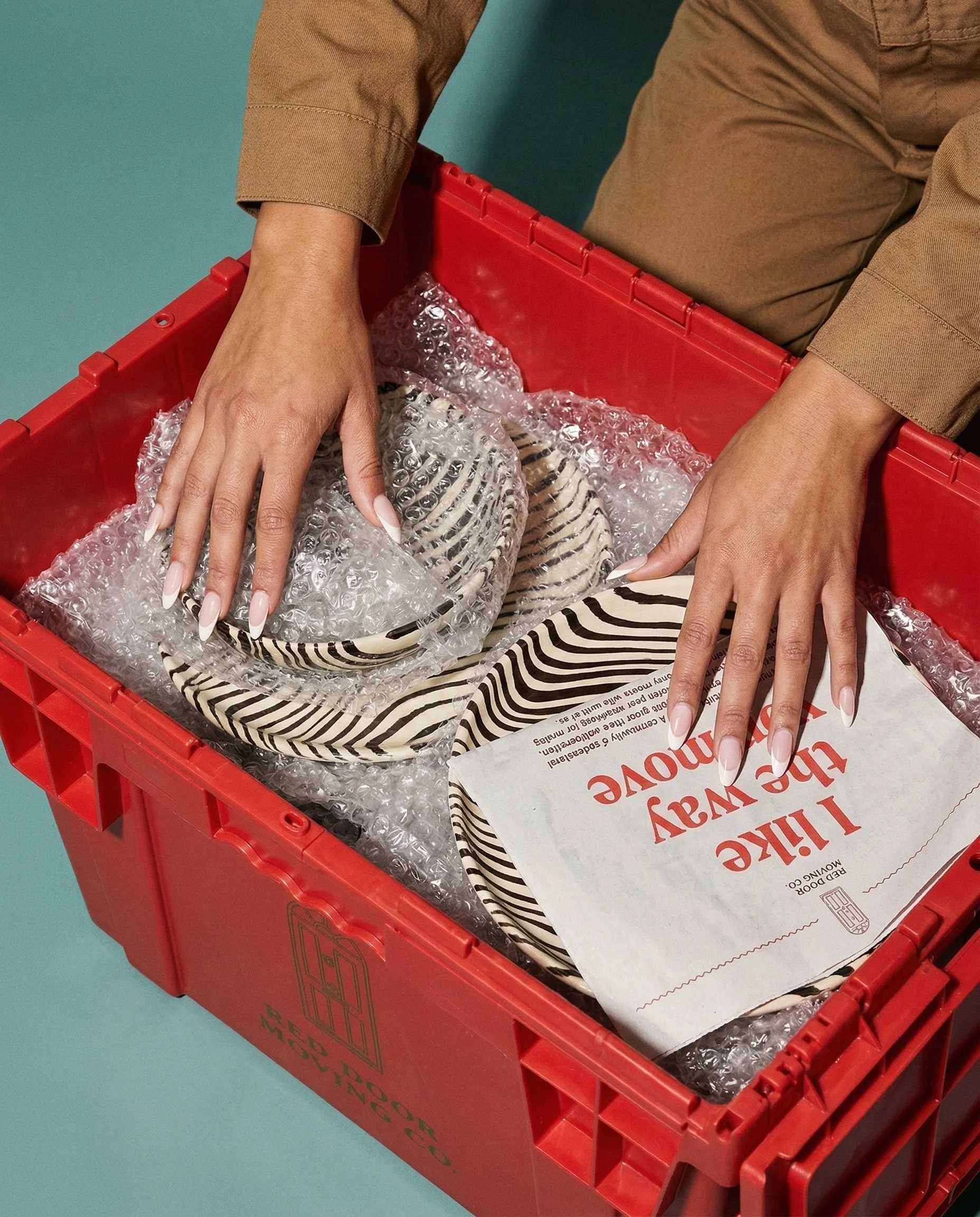 Person opening a red plastic shipping crate containing striped plates and bowls, with bubble wrap for protection, on a teal surface.