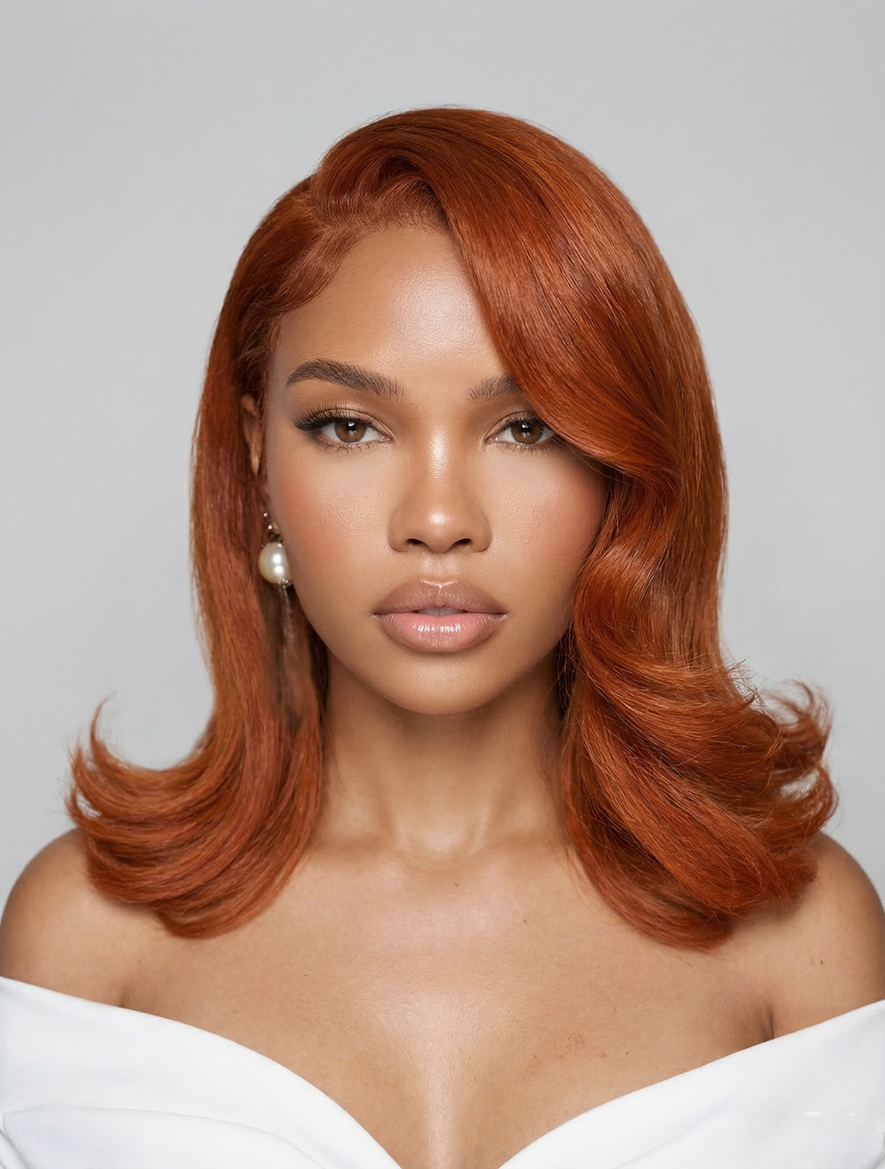 A woman with medium brown skin and shoulder-length wavy red hair, wearing pearl earrings and a white off-the-shoulder top, against a plain light gray background.