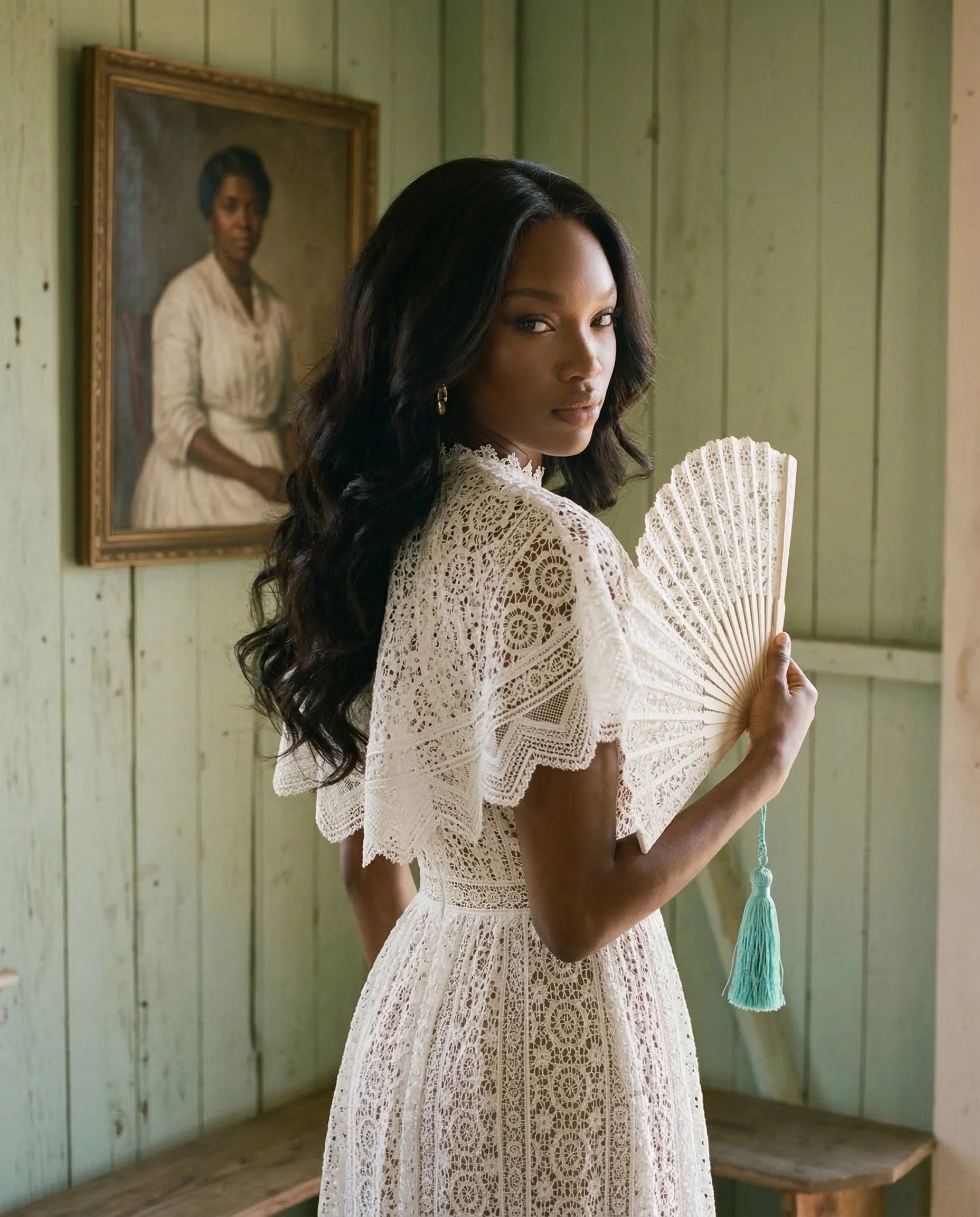 A woman in a lace dress holding a fan stands in a room with green wooden walls and a portrait of a woman in period clothing hanging behind her. Studio Abdul ai campaign image 