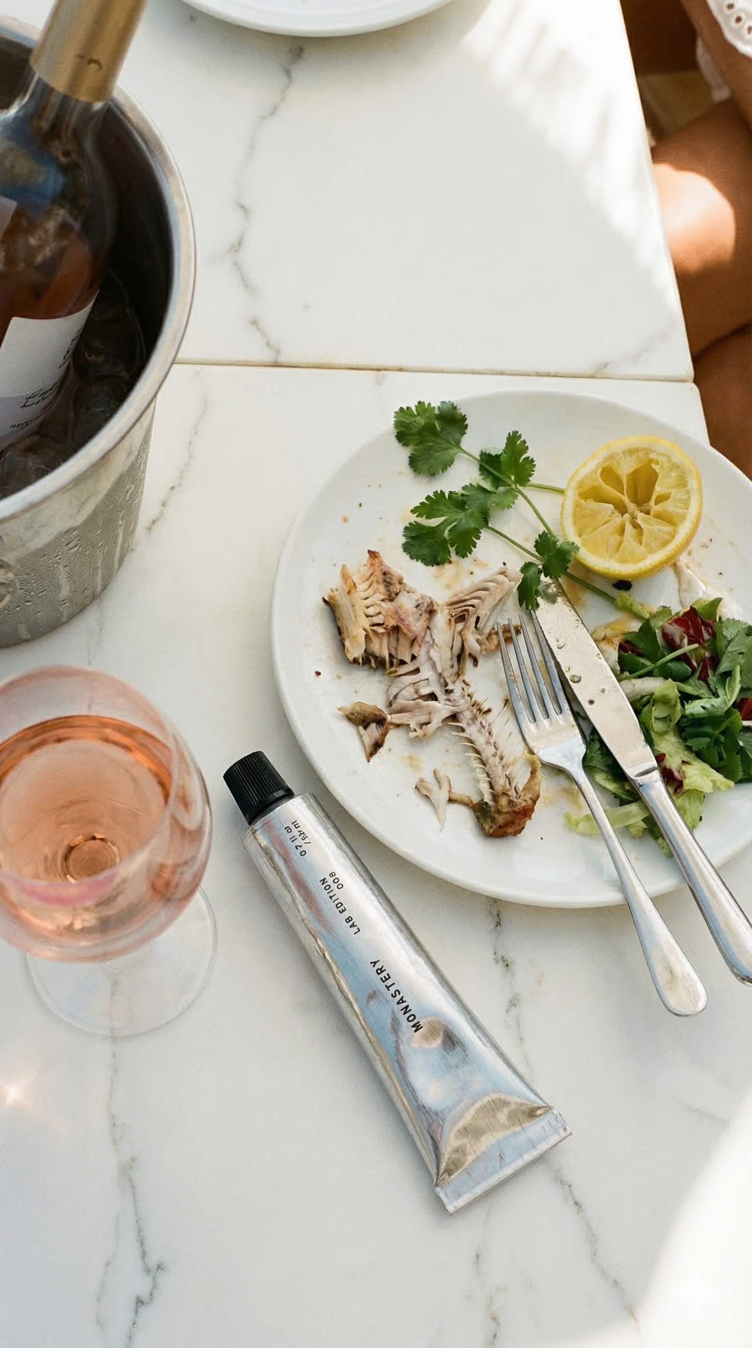 A partially eaten fish fillet with salad, lemon, and cilantro on a white plate, a glass of rosé wine, a tube of hand cream, and a metal ice bucket on a white marble table.
