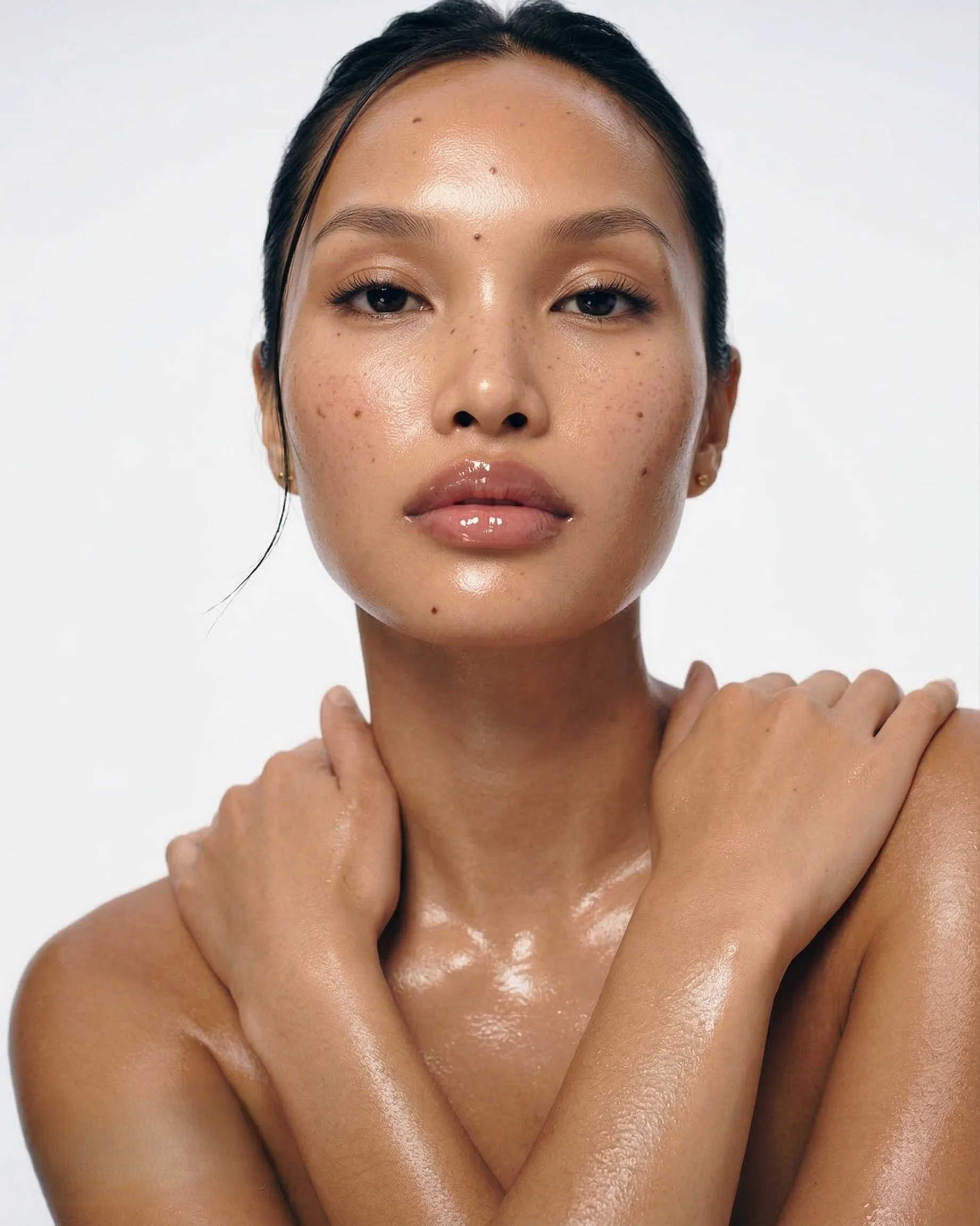 Close-up of a woman with wet skin, dark hair, and natural makeup, posing with hands crossed over shoulders against a plain white background.