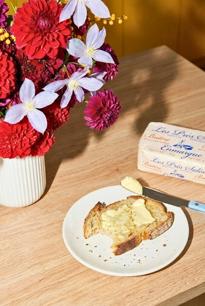 A bouquet of red, purple, and white flowers in a white ribbed vase on a wooden table, with a slice of bread with butter on a white plate, a butter knife, and a box of butter nearby.