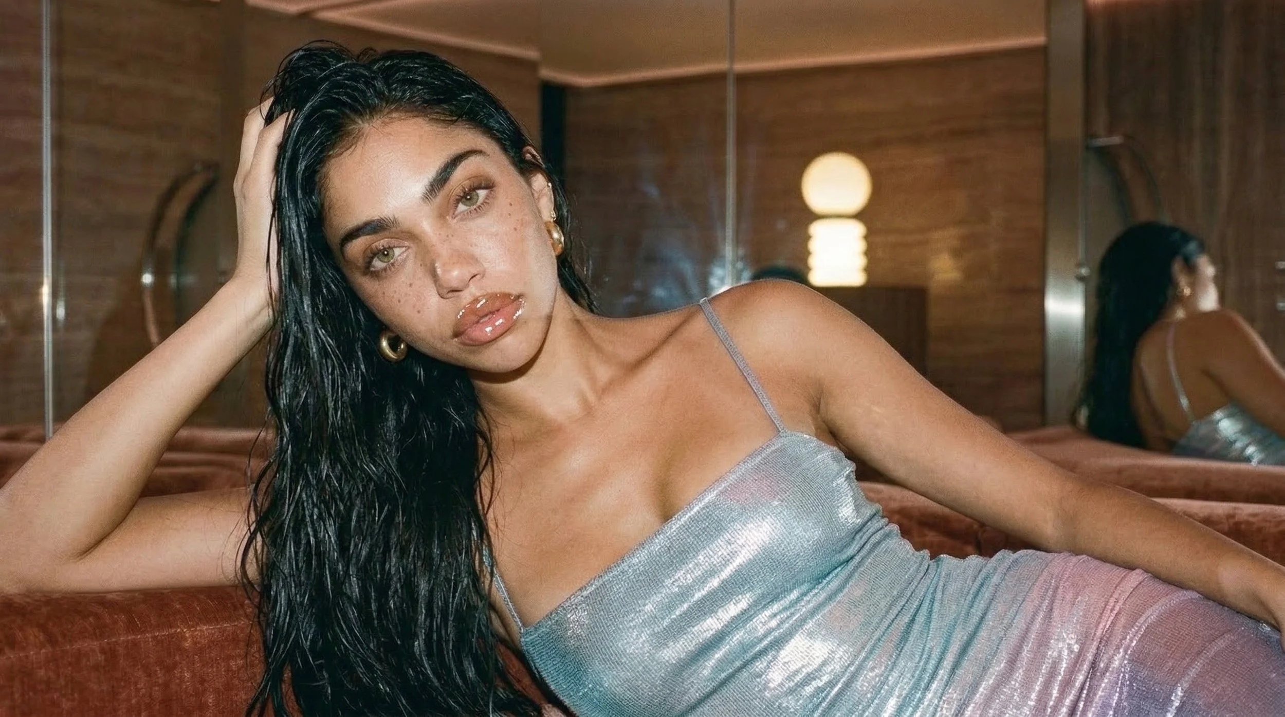 Woman with dark hair and light skin lying on a bed, wearing a shiny silver dress, with a mirror and warm lighting in the background.
