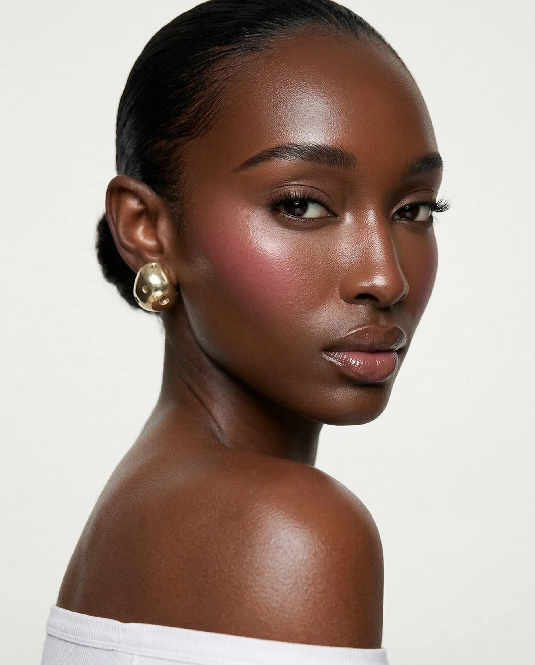 Close-up portrait of a Black woman with smooth skin, short black hair, subtle makeup, and gold hoop earrings, against a plain white background.