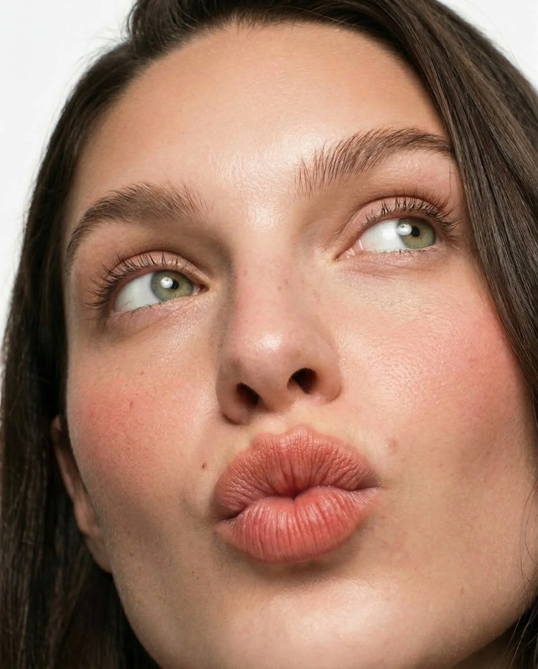 Close-up of a woman's face with her lips puckered, looking up and to the side, showing her green eyes and well-groomed eyebrows.