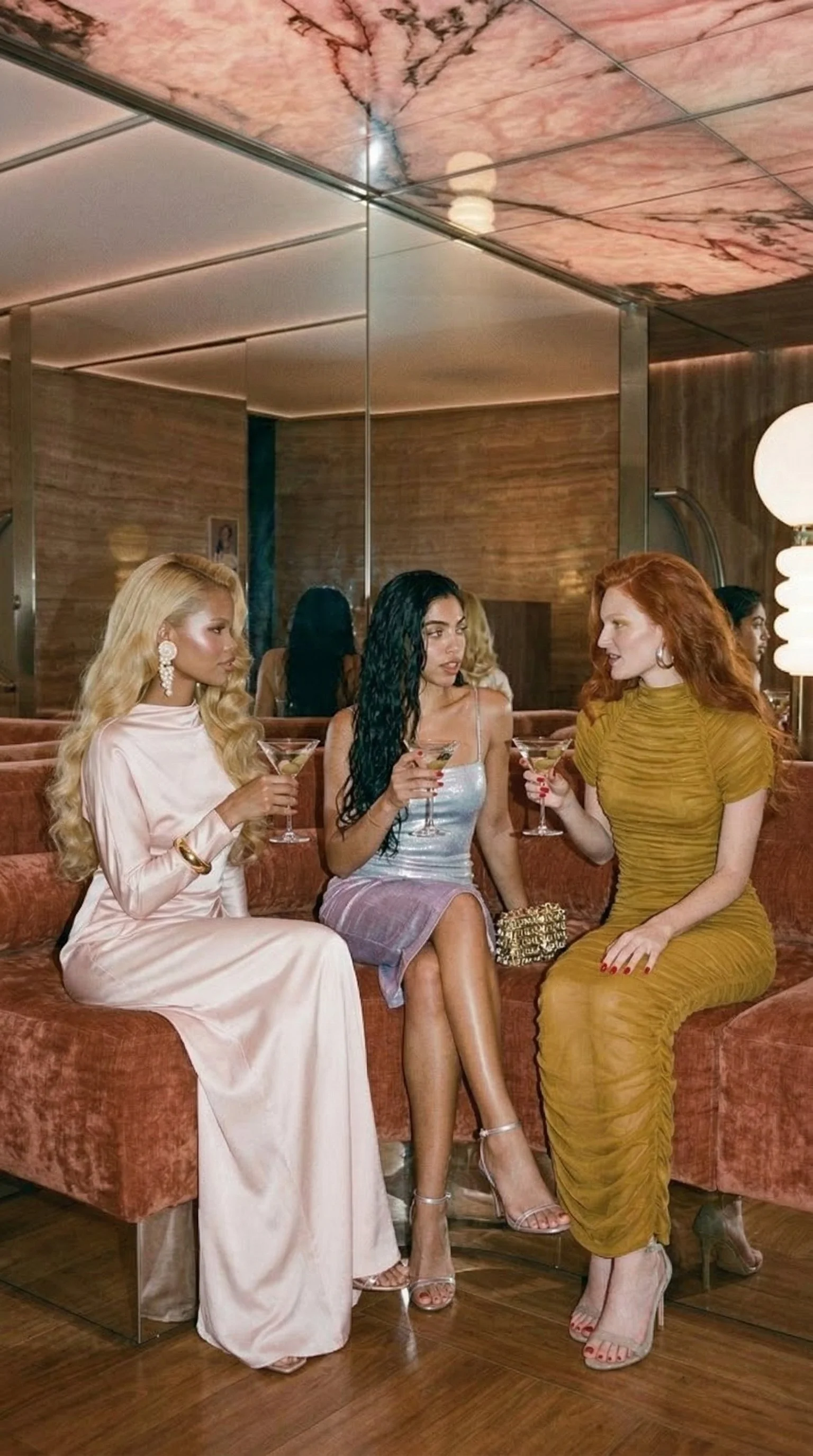 Three women in elegant dresses sitting on a pink velvet couch, holding martini glasses, having a conversation in a stylish restaurant or lounge with mirrored walls and warm lighting.