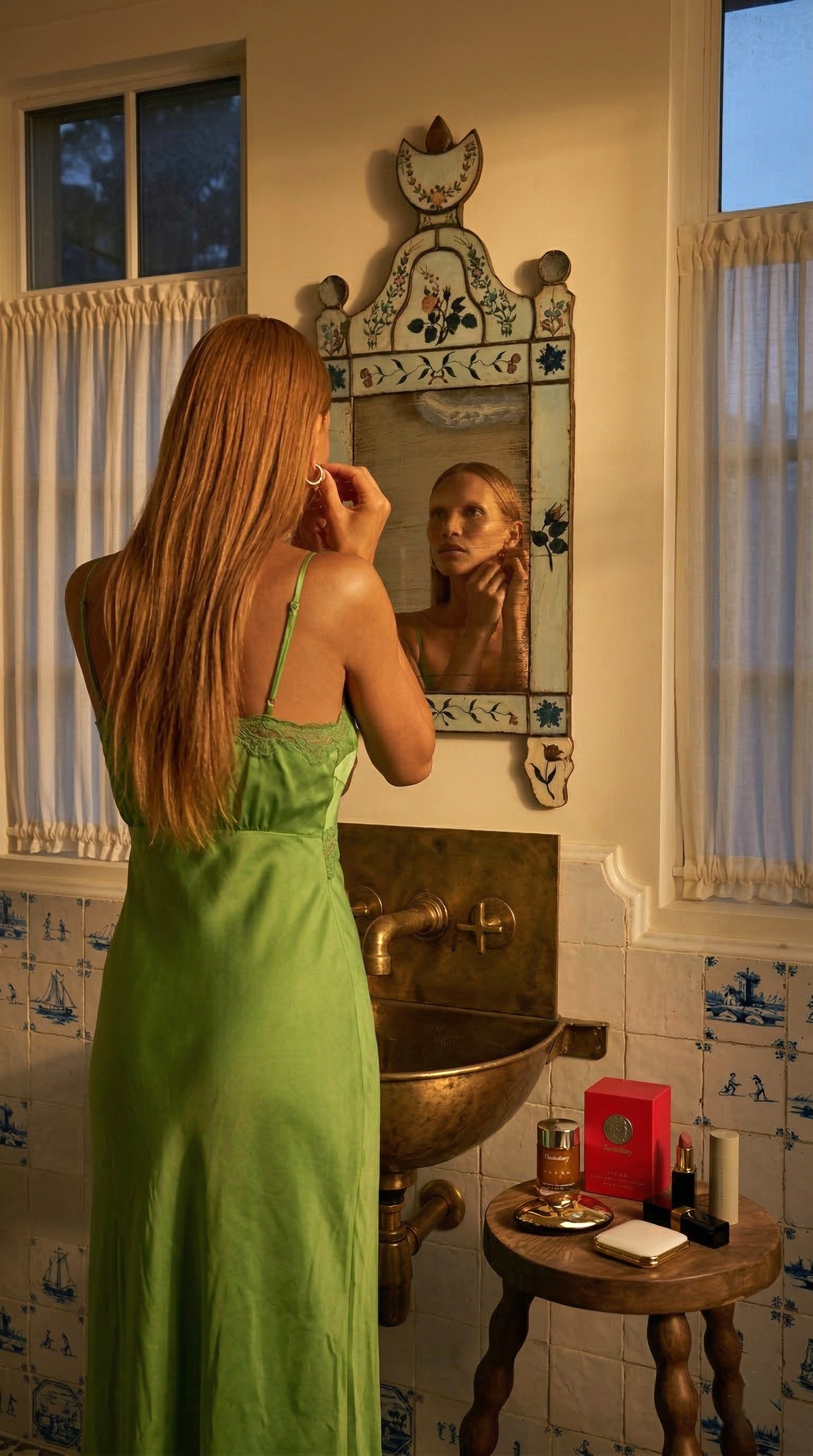 A woman with long red hair stands in front of a small bathroom mirror, applying makeup. She is wearing a green satin dress with thin straps. The bathroom has a vintage style with blue and white tiles, a brass sink, and a small wooden table holding ma