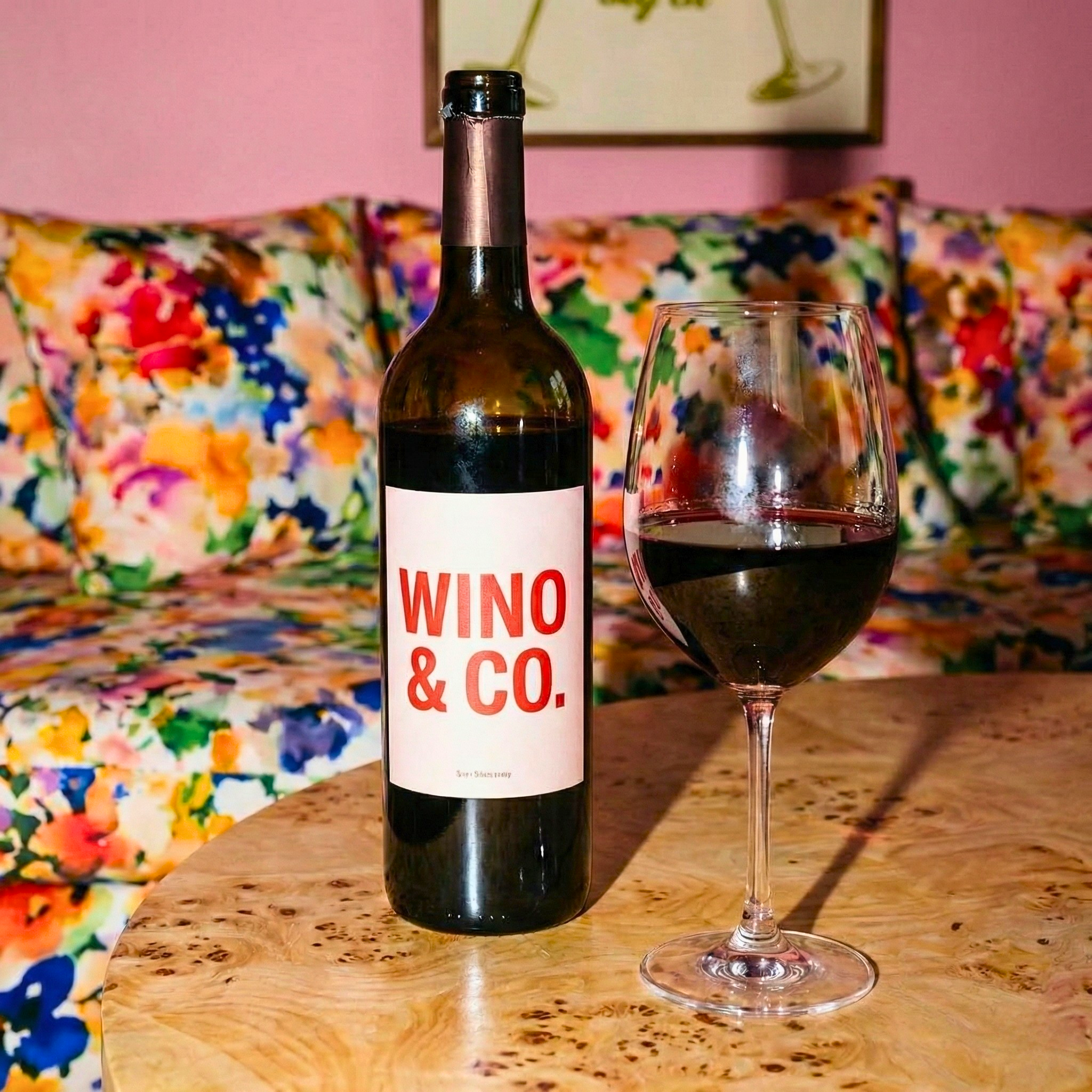 A bottle of red wine labeled 'Wino & Co.' and a filled wine glass on a wooden table in front of a colorful floral-patterned sofa.