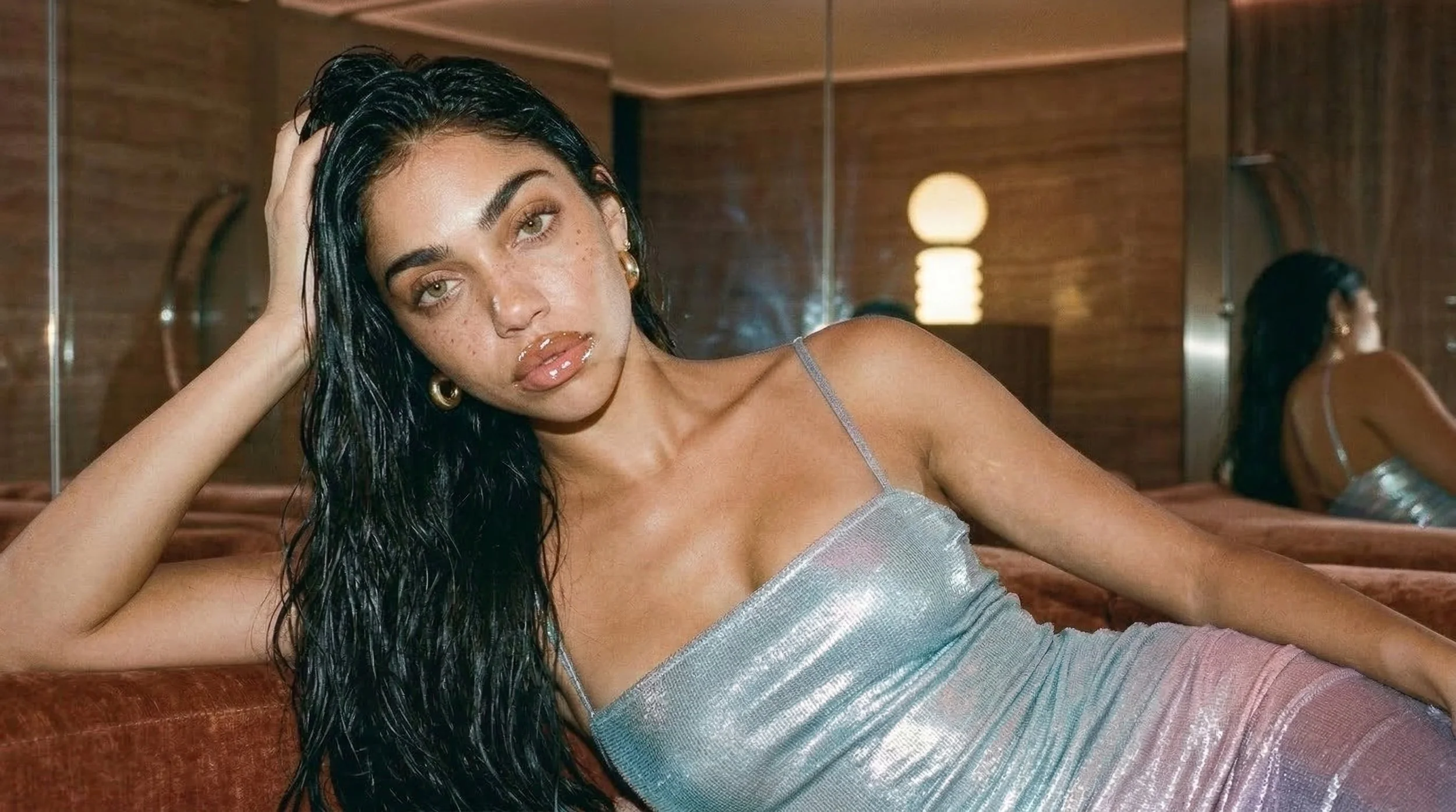 A woman with dark, wet hair lounging on a brown sofa, wearing a shimmering silver top and looking at the camera, with reflections visible in a mirror behind her.