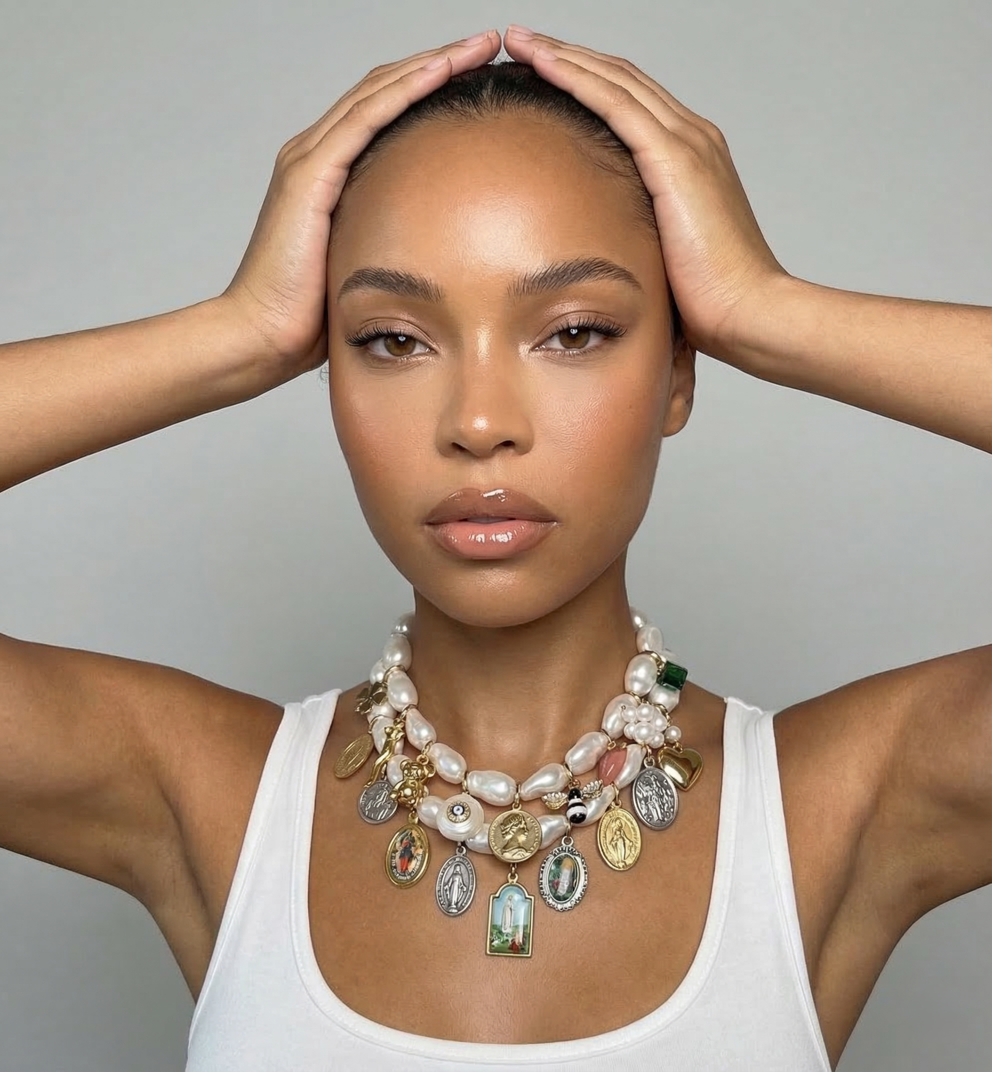 A woman with tan skin, wearing a white tank top and layered jewelry, holding her head with both hands, looking directly at the camera.