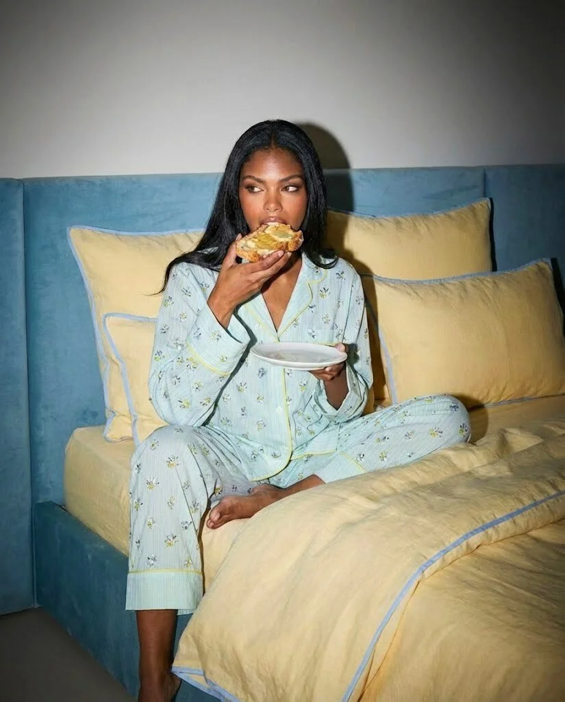 A woman sitting on a bed in pajamas, eating a slice of pizza from a plate.