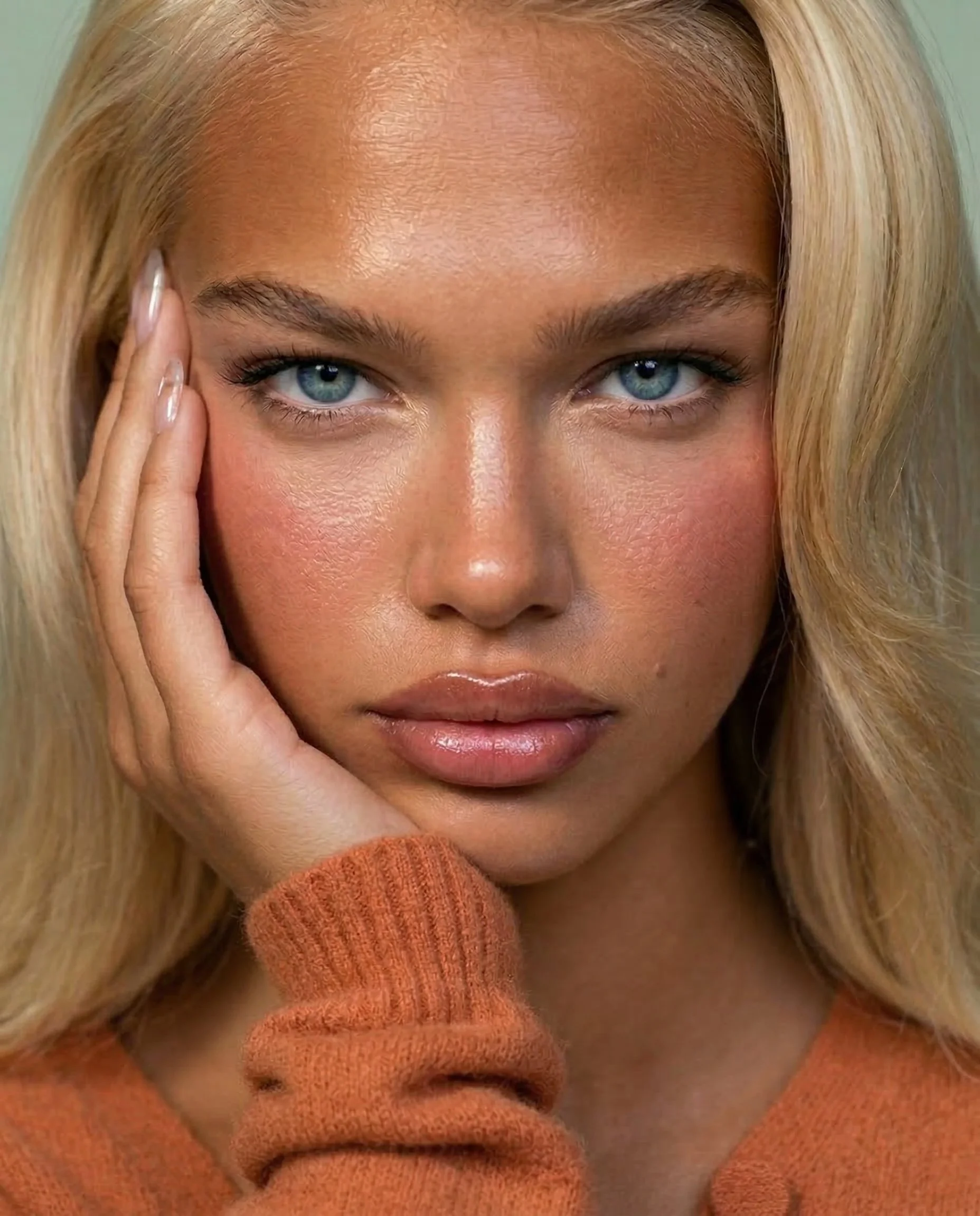 Close-up of a woman with blonde hair, blue eyes, and fair skin, wearing an orange sweater, looking directly at the camera with her hand resting on the side of her face.