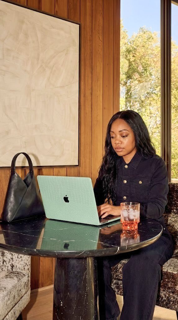 A young woman with black hair, wearing a black denim jacket, working on a light green laptop at a round black table in a cozy, wood-paneled room with a large window showing trees outside. There's a black handbag on the table and a glass of pink beverage.