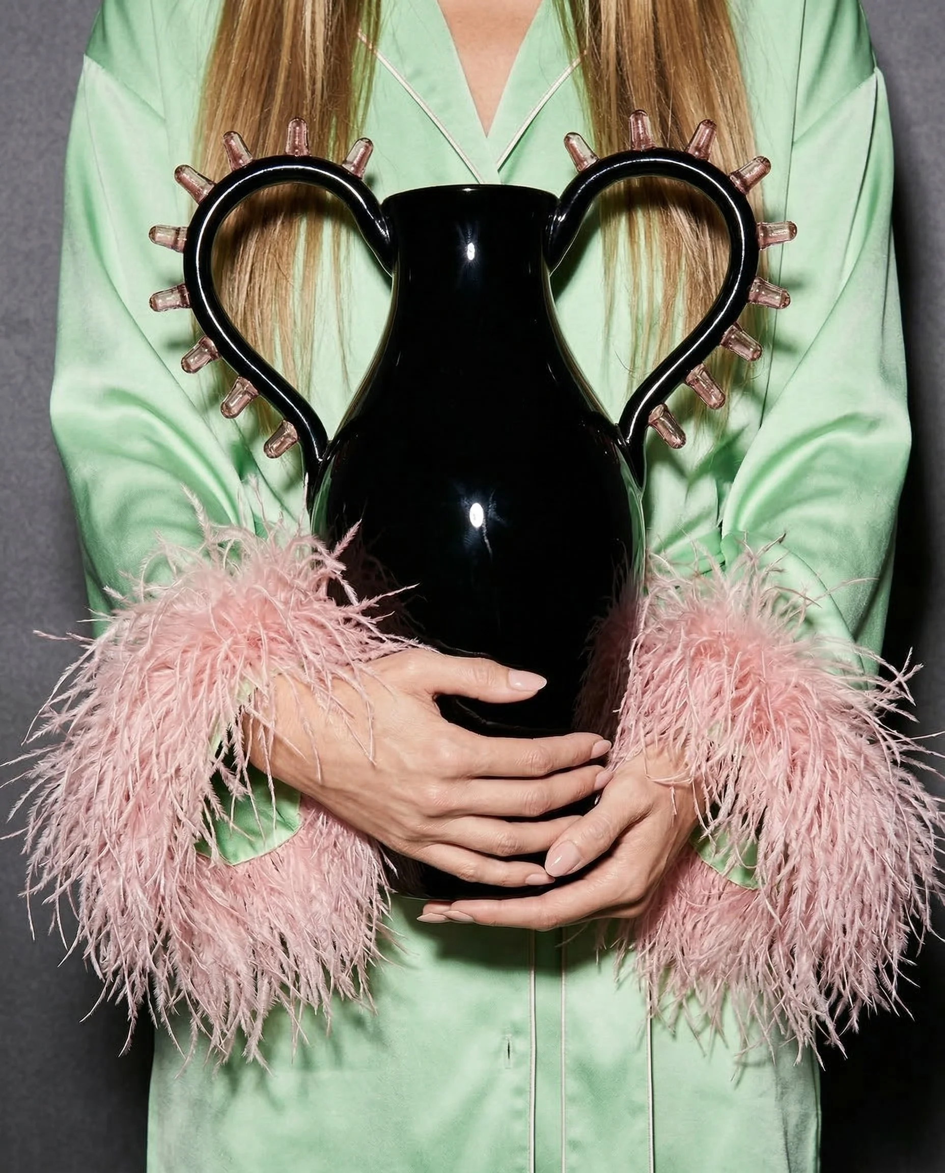 Person wearing a green satin robe with pink feather cuffs, holding a black vase with heart-shaped handles and pink tipped spikes.