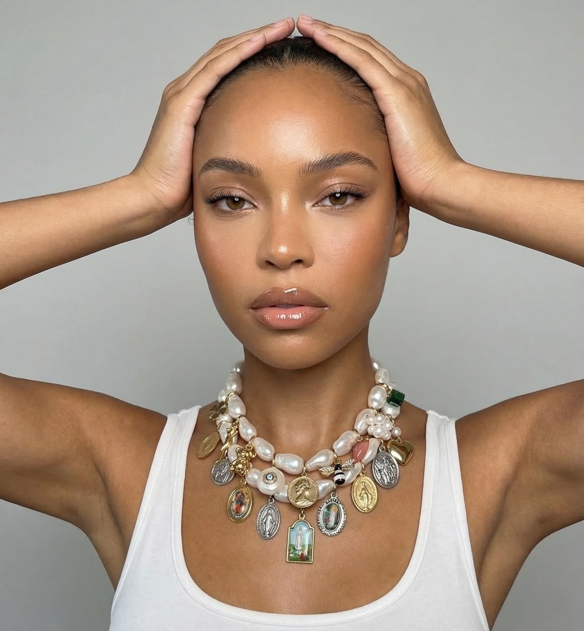 A woman with light brown skin and brown eyes holding her hair back, wearing a white tank top and layered necklaces with religious and decorative pendants.