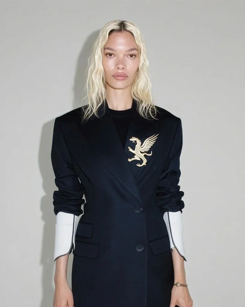 A woman with blonde, wavy hair wearing a black blazer with a gold winged lion emblem on the left side and white accents on the sleeves, standing against a plain light gray background.