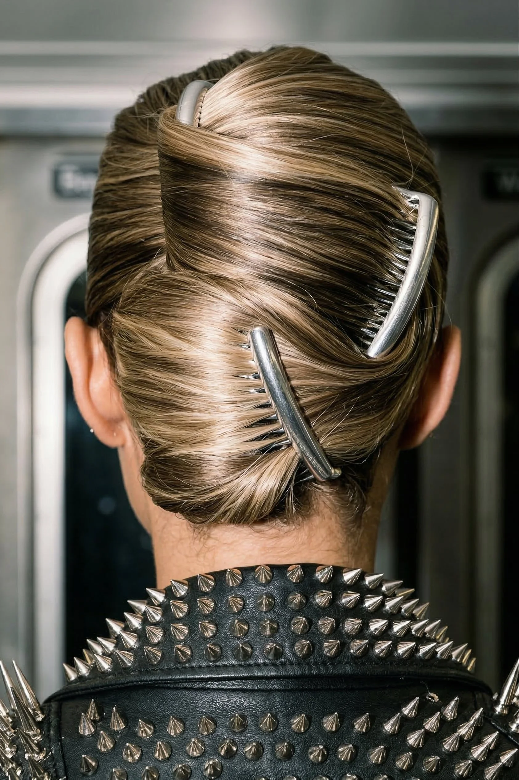 A person with blonde hair in a bun, secured with large metallic hair clips, wearing a black jacket with metal spikes, standing in front of a metallic door.