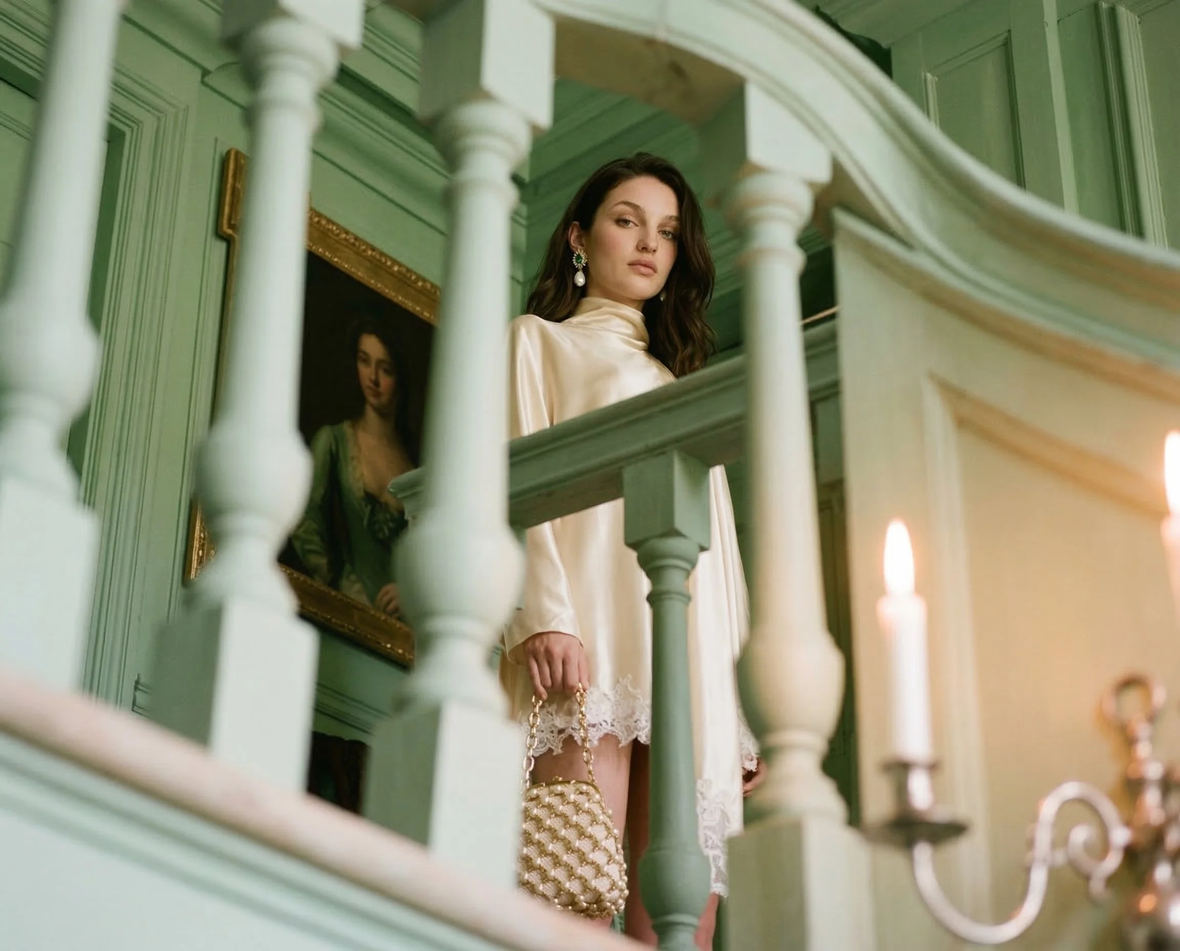 A woman with dark hair wearing a cream-colored satin dress, holding a quilted purse, standing on a staircase with green walls and a framed portrait behind her. Studio Abdul ai campaign image 